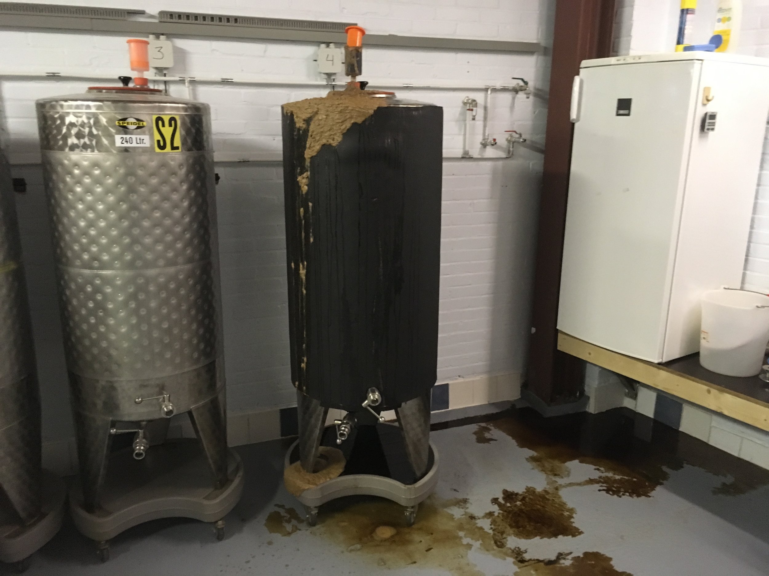 In een ruimte staan twee grote roestvrijstalen tanks en een zwarte houten standaard met eroverheen een ander materiaal dat eruitziet als compost of mest, met een druppelende vloeistof. Er ligt afval op de vloer onder de standaard.