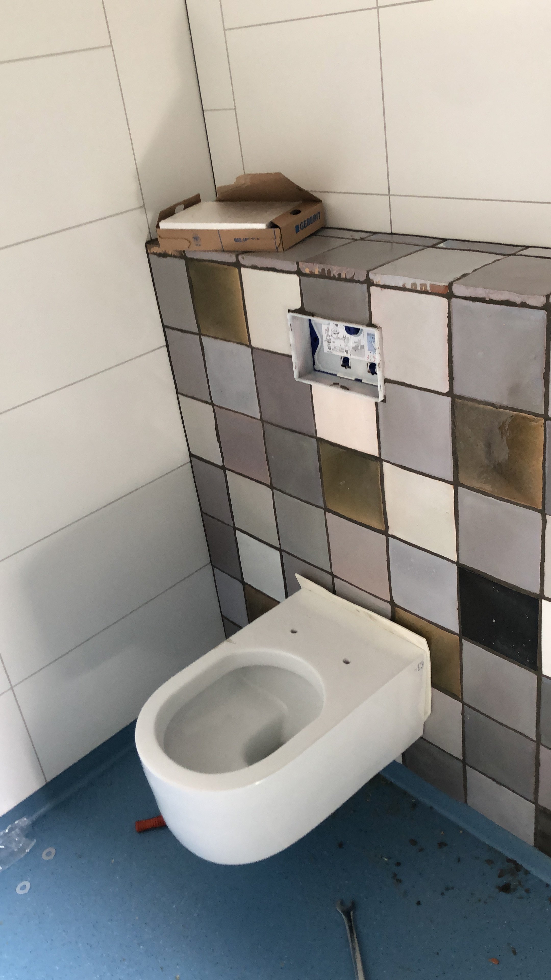 Onvoltooide toiletruimte met een losstaand toilet en een muurbuffer met keramische tegels. Er liggen gereedschap en bouwstof op de vloer.