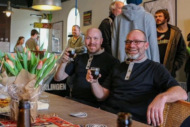 Twee mannen zitten aan een tafel in een café of restaurant, glimlachend en met een glas in hun hand. Ze lijken plezier te hebben. Op de tafel staan tulpen, drankjes en snacks, en er zijn andere mensen op de achtergrond.