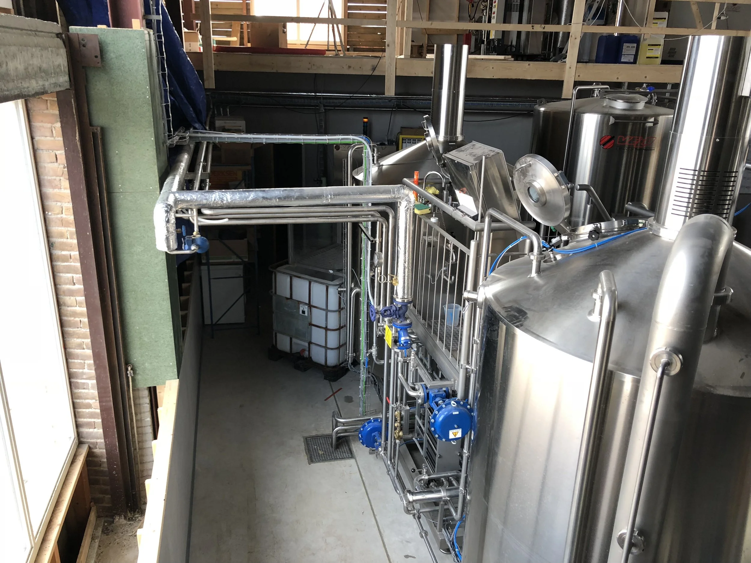 Binnenkant van een industriële installatie met metalen pijpen, tank en controlepanelen, gesitueerd in een ruimte met houten en bakstenen muren.