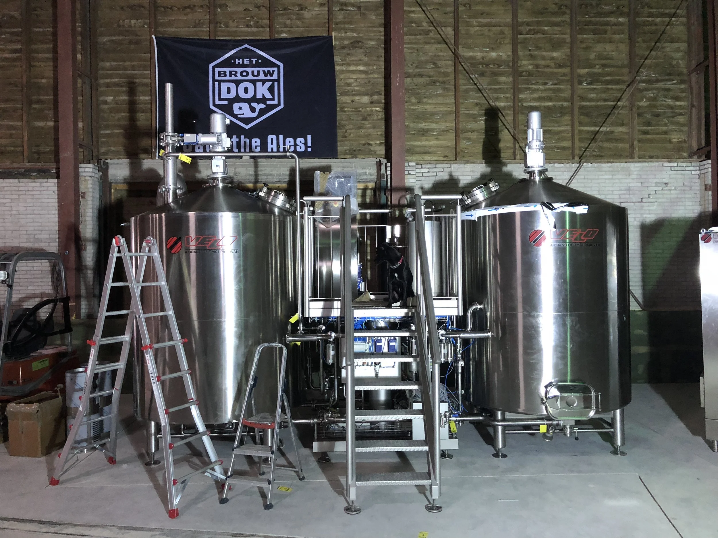 Stalen tanks in een fabriekshal met een blauwe banner met de tekst 'HET BROUW DOK' en 'voor de Ales!'