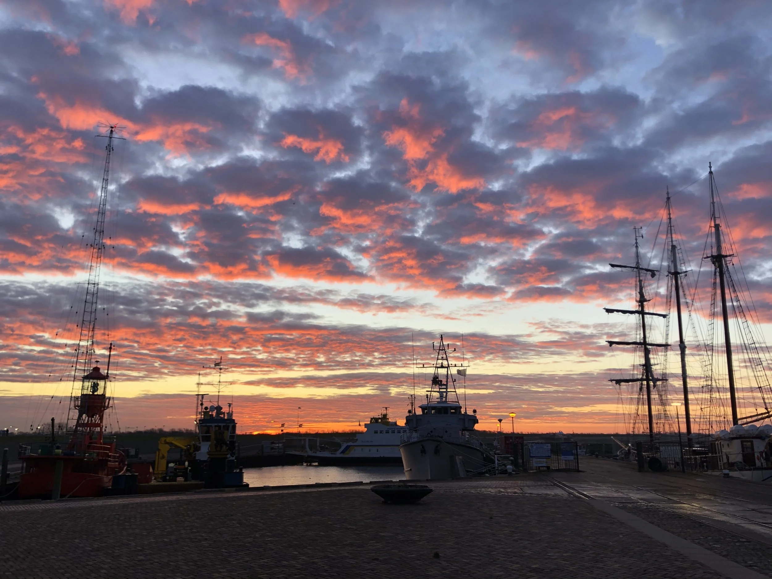 Kade in de haven bij zonsondergang met schepen en een kleurrijke hemel met roze en blauwe wolken.