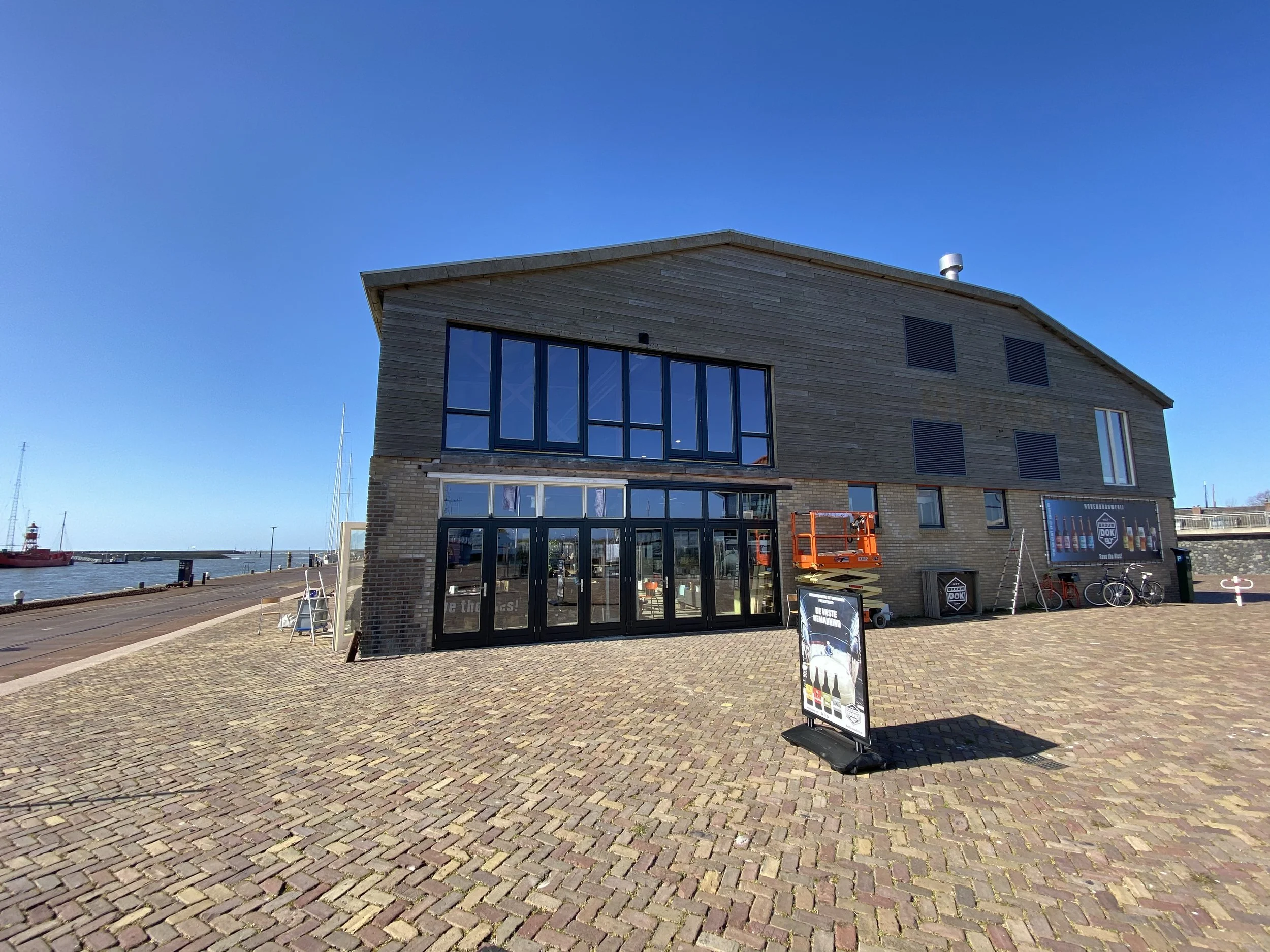 Een modern gebouw met grote ramen bij de haven, een verlichte gevel, een informatiebord en fietsen op de voorgrond, met een blauwe lucht en water op de achtergrond.