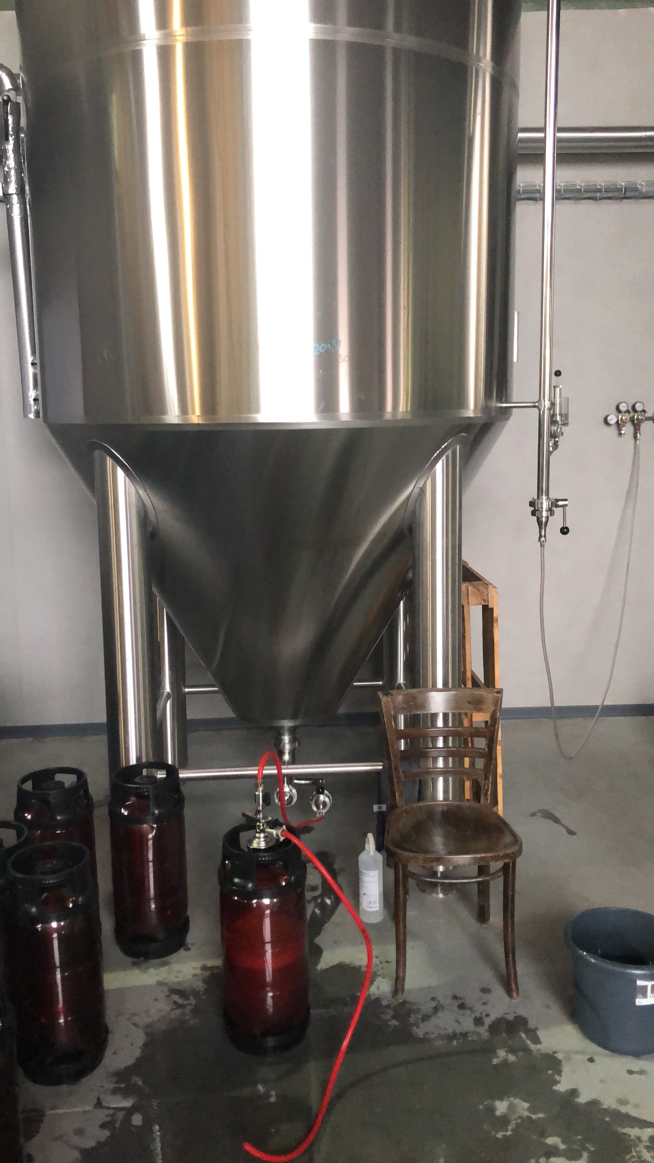 Een grote roestvrijstalen tank met een conische onderkant, gebruikt voor het fermenteren of opslaan van vloeistoffen, staat in een industriële omgeving. Er staan lege bierflesjes, een stoel en enkele hulpmiddelen op de vloer eronder en ernaast.