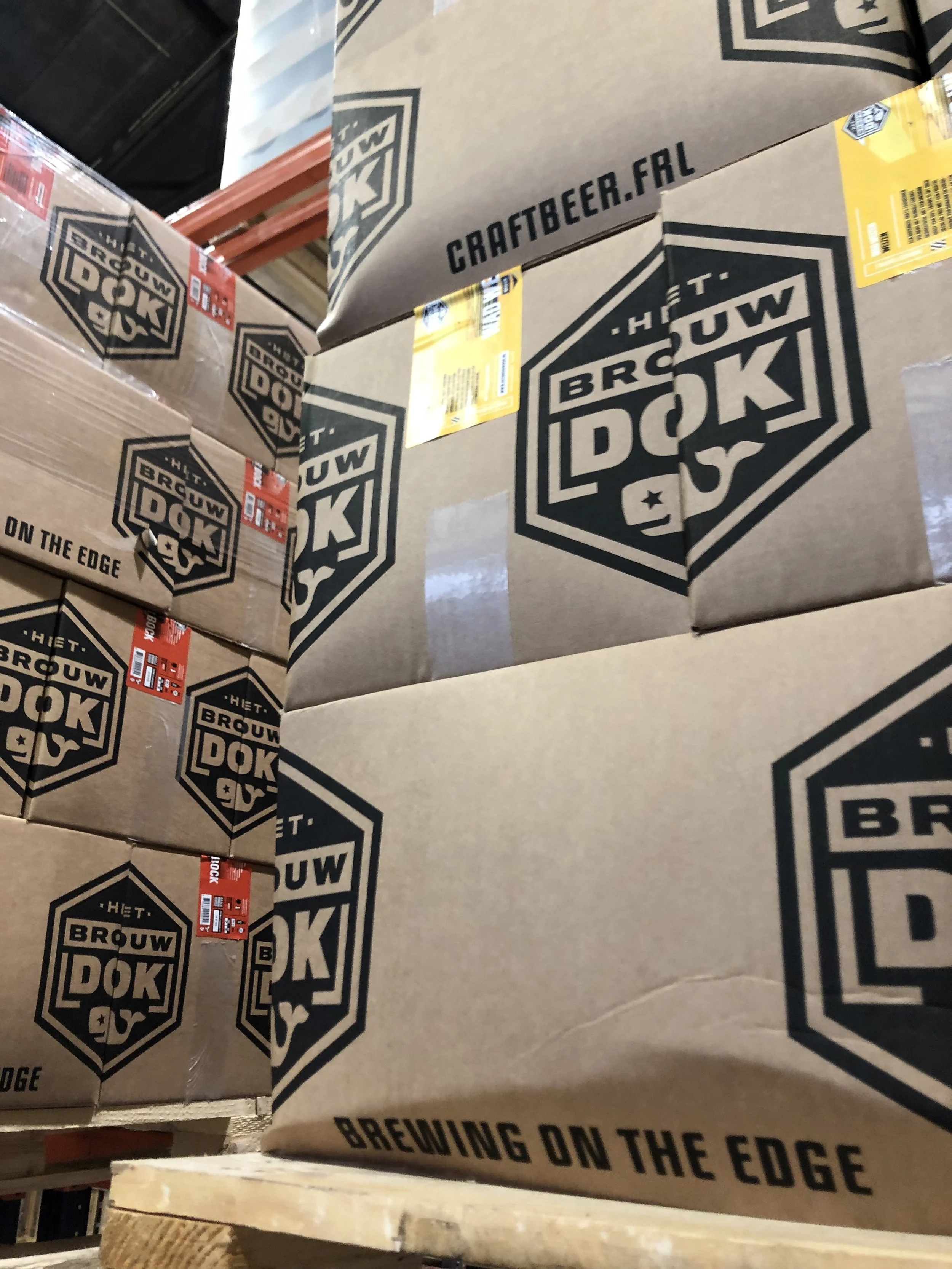 Daar liggen kartonnen dozen met het logo van Brouw DOK, ze zijn op een pallet in een winkel of magazijn.