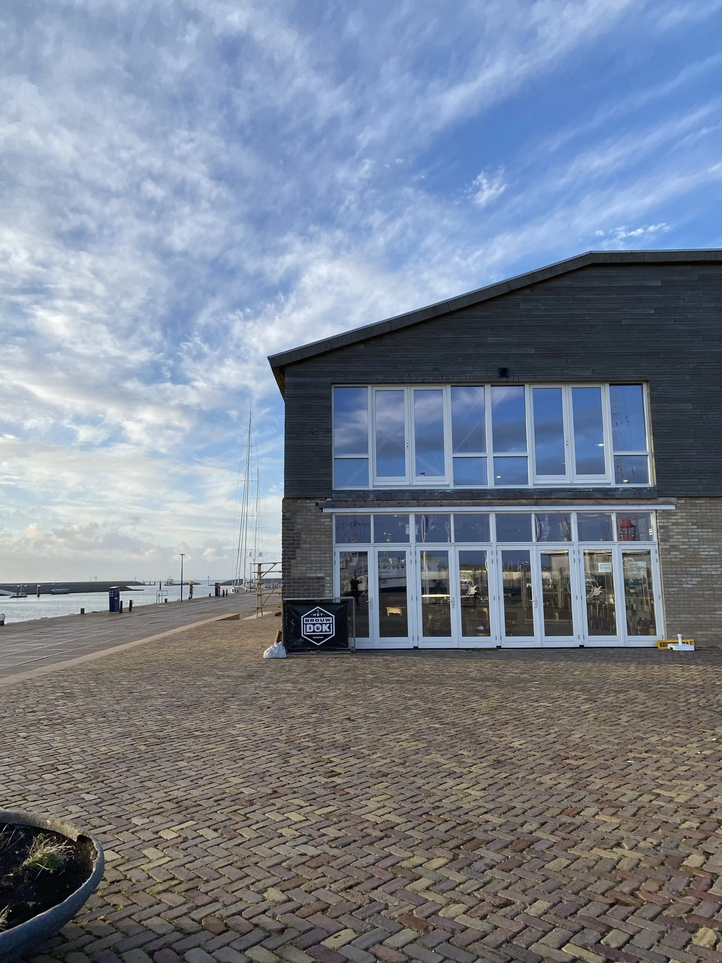 Een gebouw met grote ramen en een dak op de kade, met een zwart bord met witte tekst 'Bouw DOK' en een skyline, onder een bewolkte blauwe hemel.