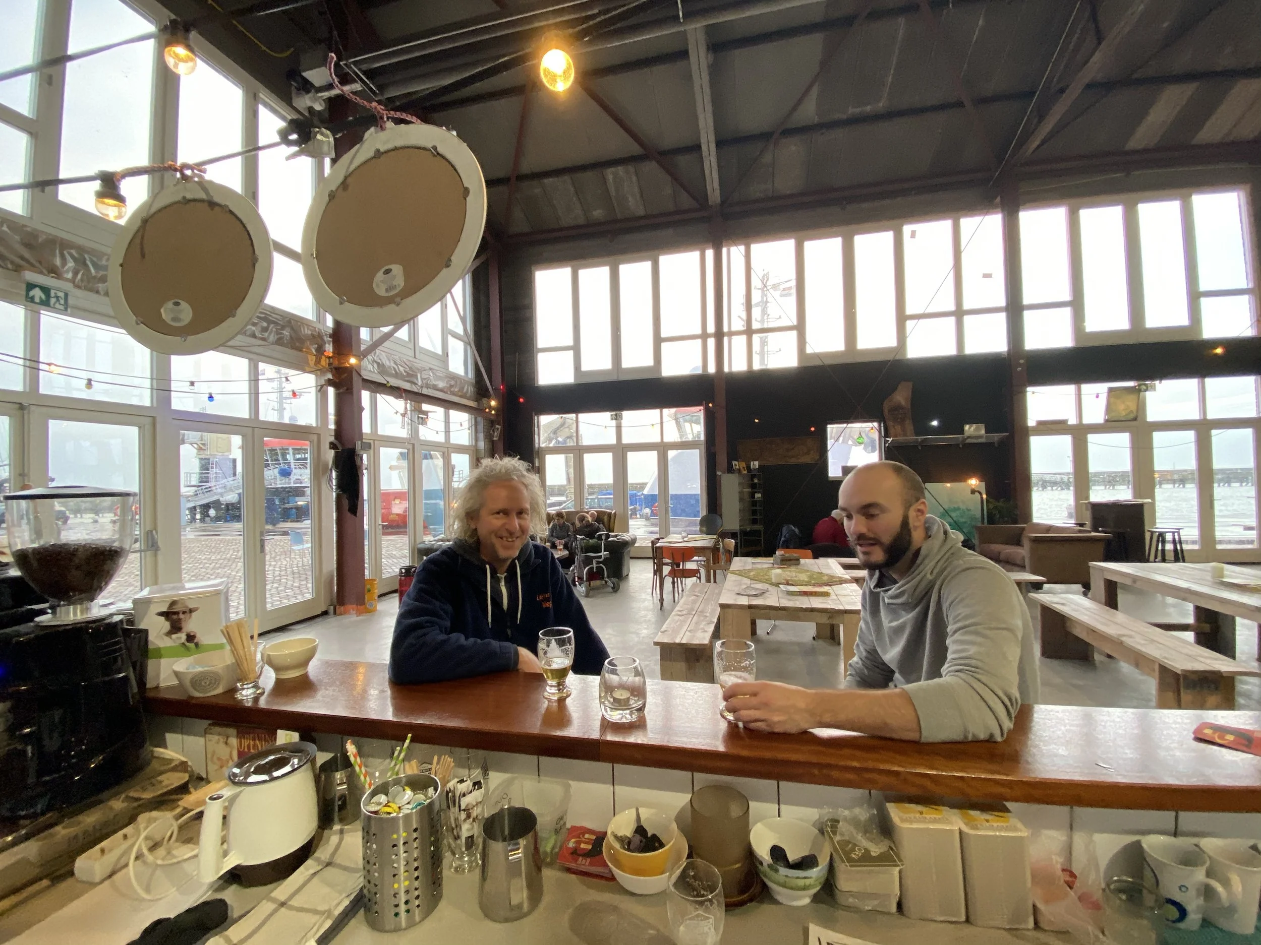 Twee mannen zitten aan een tafel in een restaurant, drinkend uit glas. Achter hen grote ramen met uitzicht op een haven met schepen.