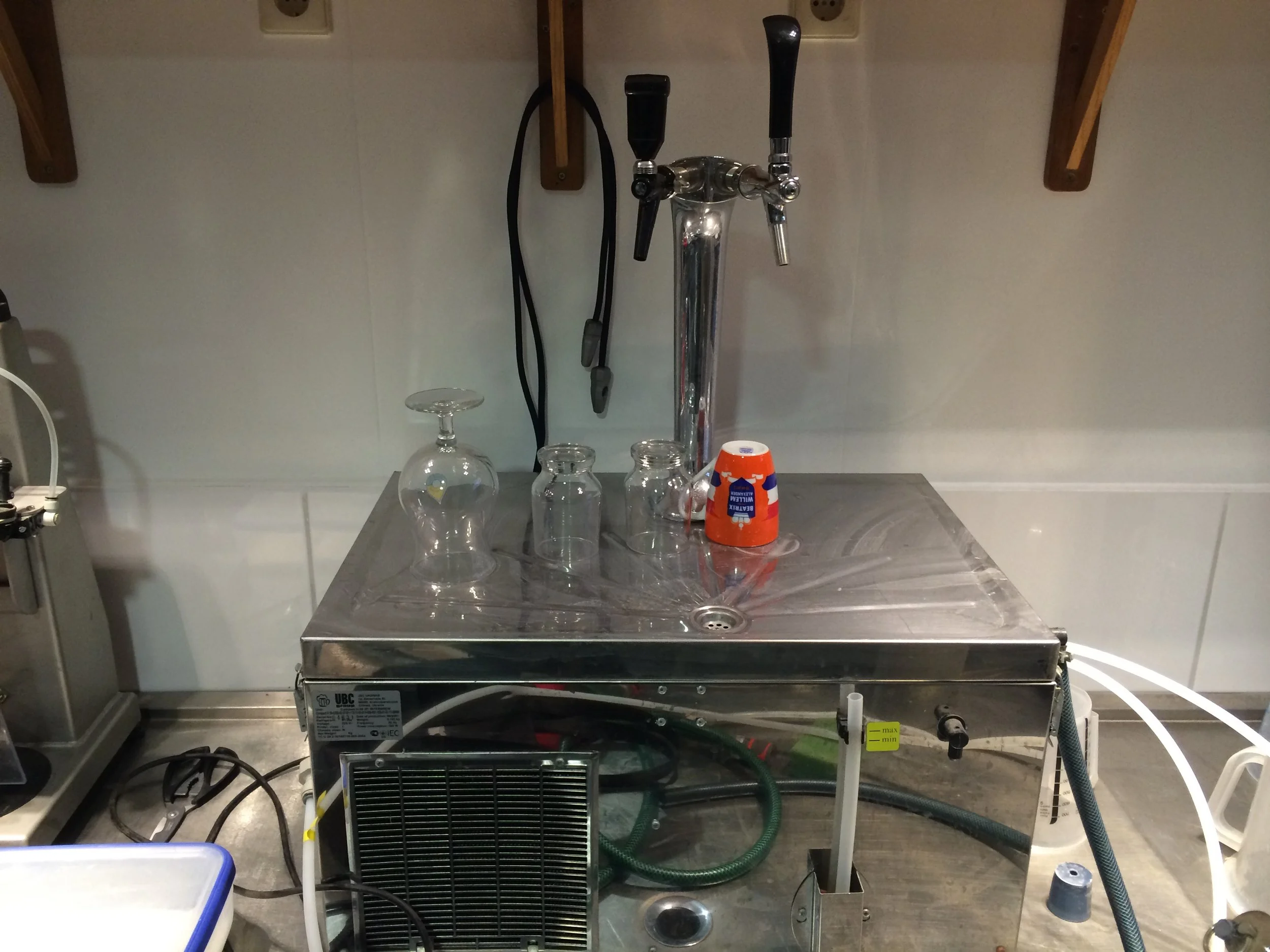 Een laboratoriumopstelling met een glasfles, vier kleine glazen bekers, een oranje bekertje met tekst, een stainless steel apparaat met hendel, een apparaat met draden en een ventilator onderin, en verschillende slangen en kabels.