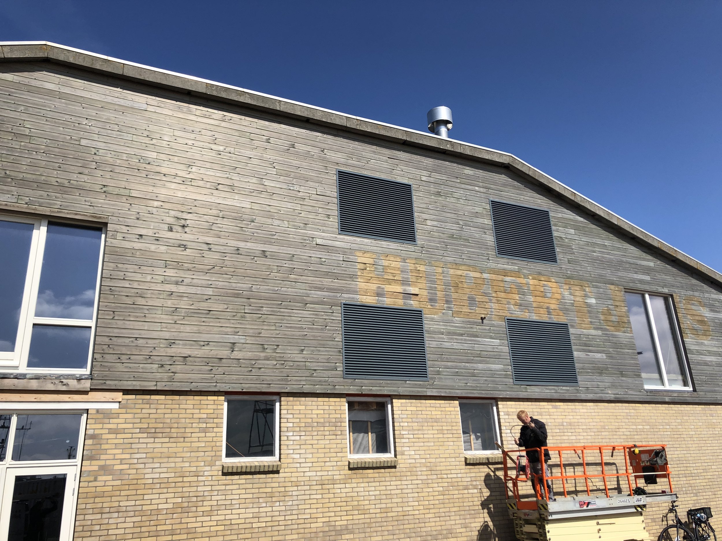 Een gebouw met een houten bovenverdieping en een onderste verdieping van bakstenen, ramen en ventilatieroosters. Een persoon staat op een steiger vlakbij het gebouw.