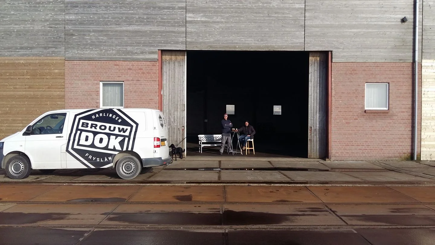 Een witte bedrijfsauto met logo 'Brouwdok Harlingen Fryslan' geparkeerd voor een oud gebouw met een grote open ingang. Twee mensen staan en zitten bij een kleine tafel binnen in de schaduw, naast een bankje. Twee kleine honden zijn aanwezig, een naas