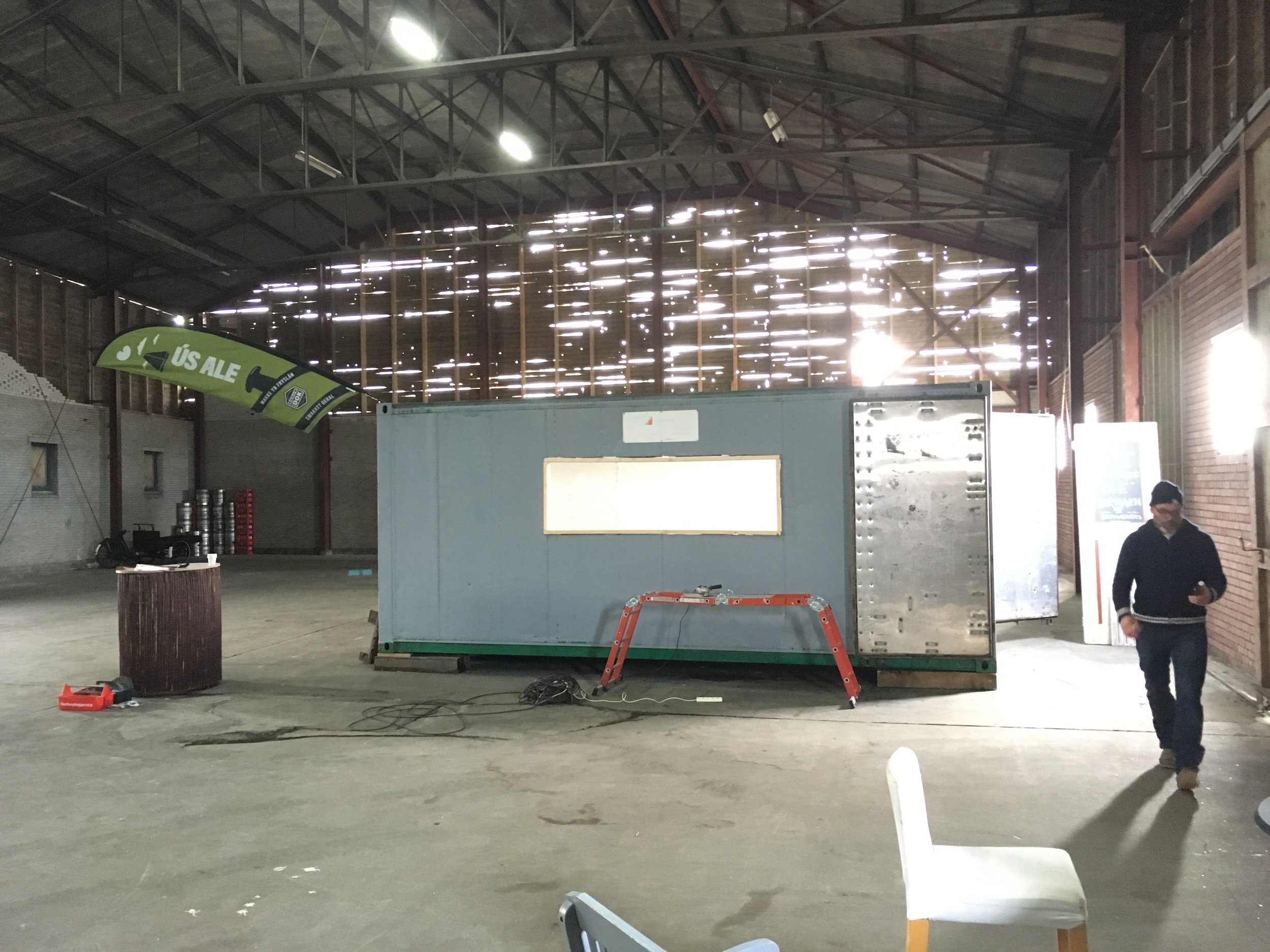 Binnenruimte van een grote loods of magazijn met constructiewerk in uitvoering. Er staat een blauwe cabine, mogelijk een mobiele kraan of werkplek, en er hangen lampen aan het plafond.