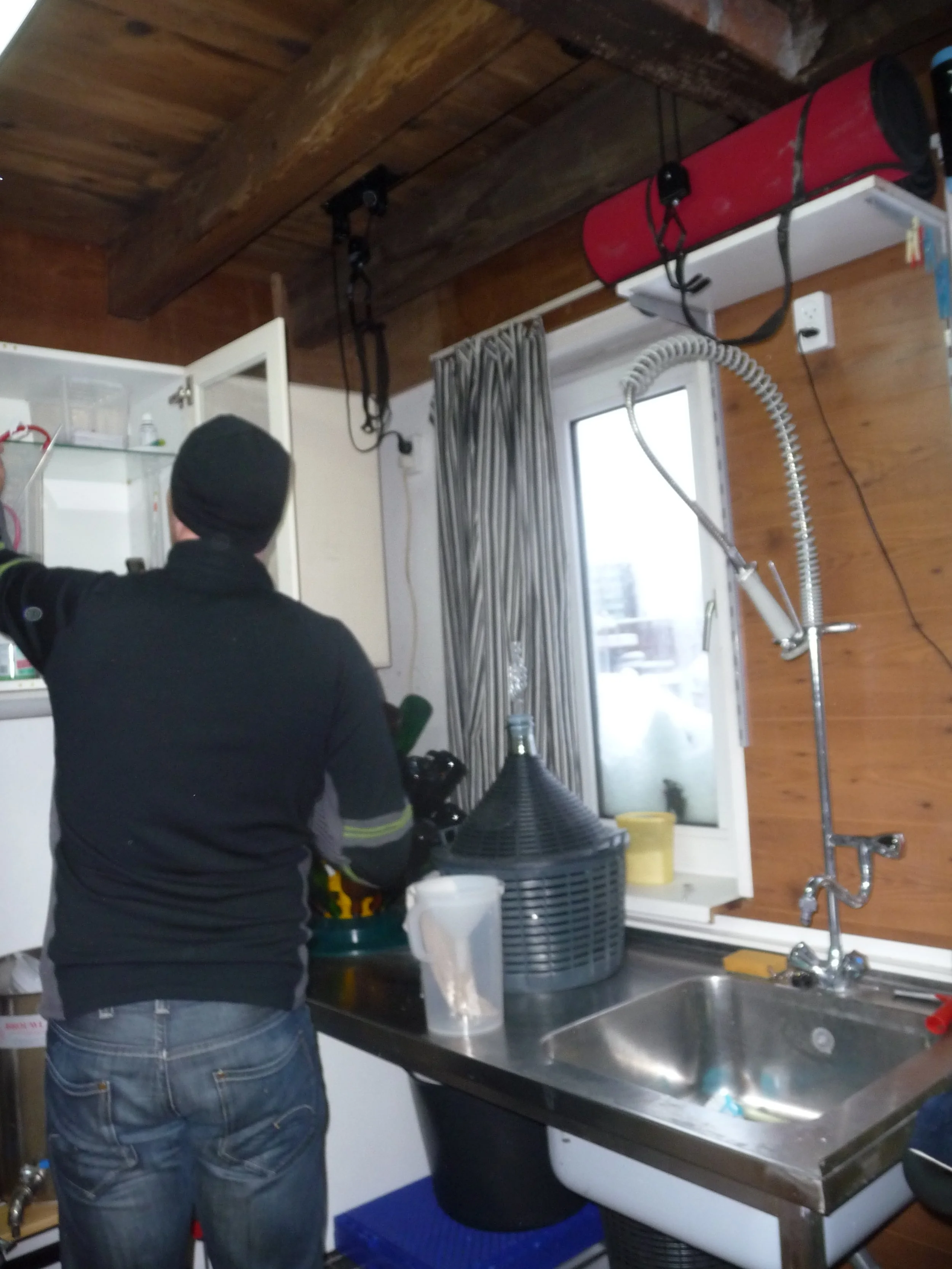 Man in zwarte jas en blauwe jeans staat in een keuken, zijn rug naar de camera, en werkt aan iets op de muur of plafond.