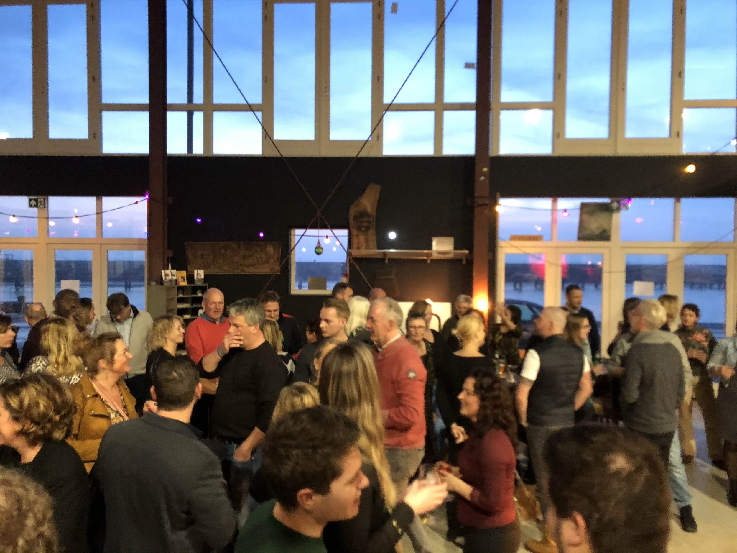 Binnenbeeld van een druk bezoekersfeest met veel mensen die praten en lachen, enkele mensen dragen casual kleding en zien vrolijk uit, met grote ramen die uitzicht bieden op een blauwe avondhemel.