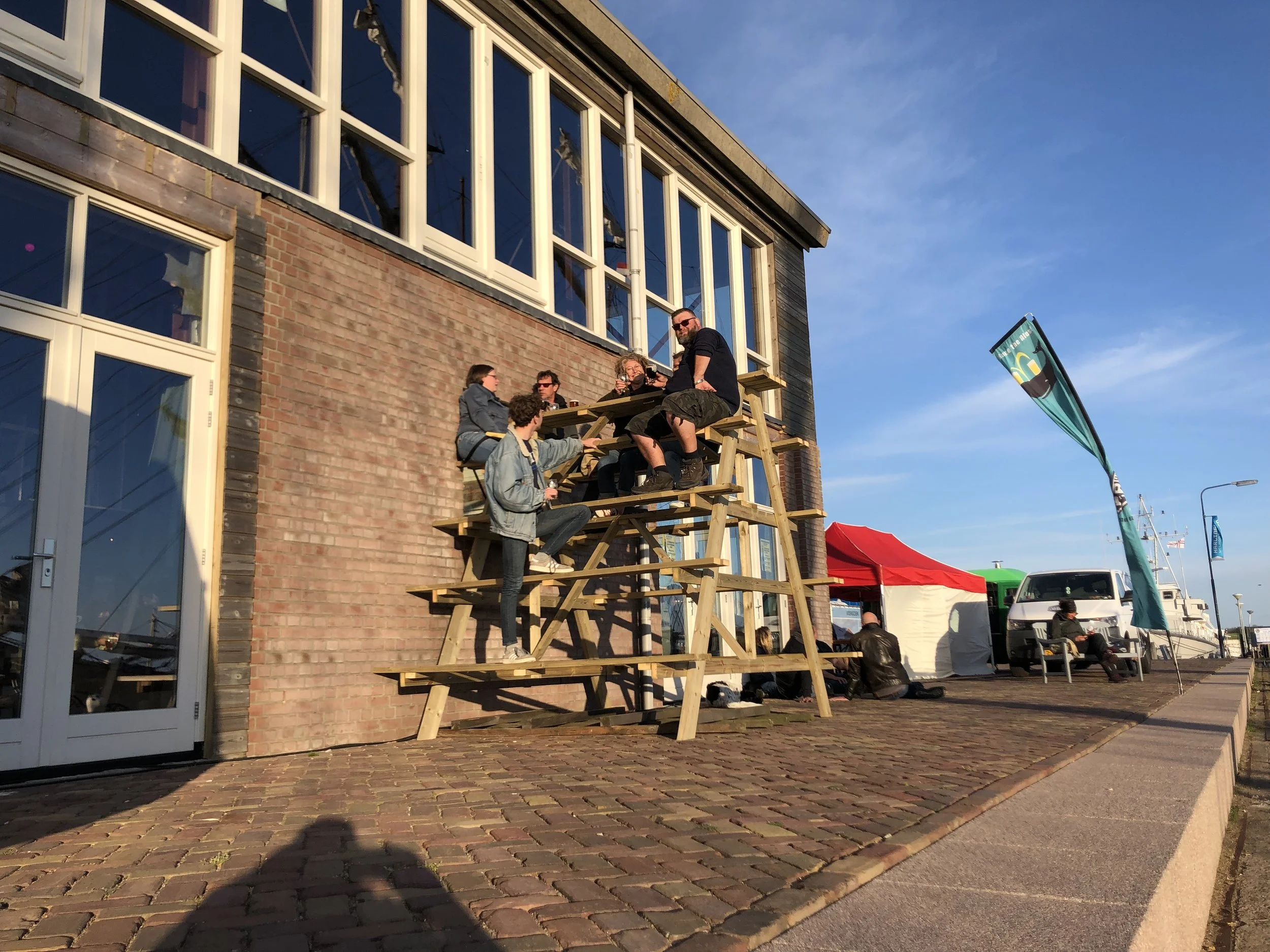 Een groep mensen zit op een houten terras, sommigen drinken en praten, terwijl ze genieten van de zon. Zij staan buiten voor een modern gebouw met grote ramen, bij een haven met boten.