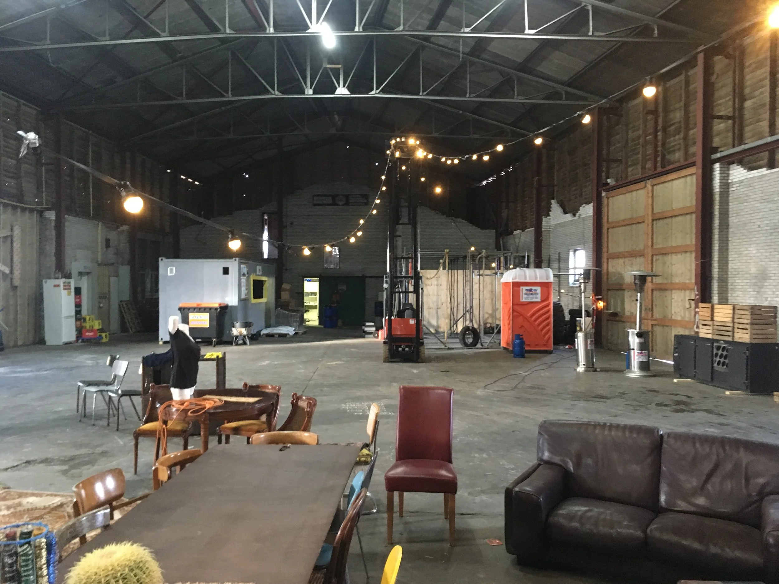 Interieur van een grote binnenruimte met een hoog, metalen, schuine dakstructuur en houten wandpanelen. Er hangen lampen en decoratieve lichtslingers. Op de vloer staan verschillende stoelen en tafels, en er zijn enkele meubels en objecten, zoals een