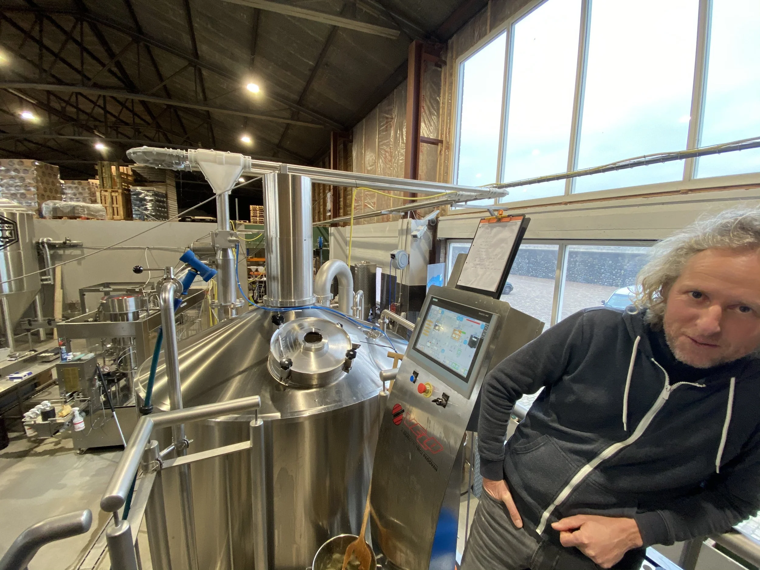 Man met grijze haar en een donkere jas staat in een laboratorium met industrieel opstelling en computerscherm, naast een grote metalen container met buizen en apparatuur.