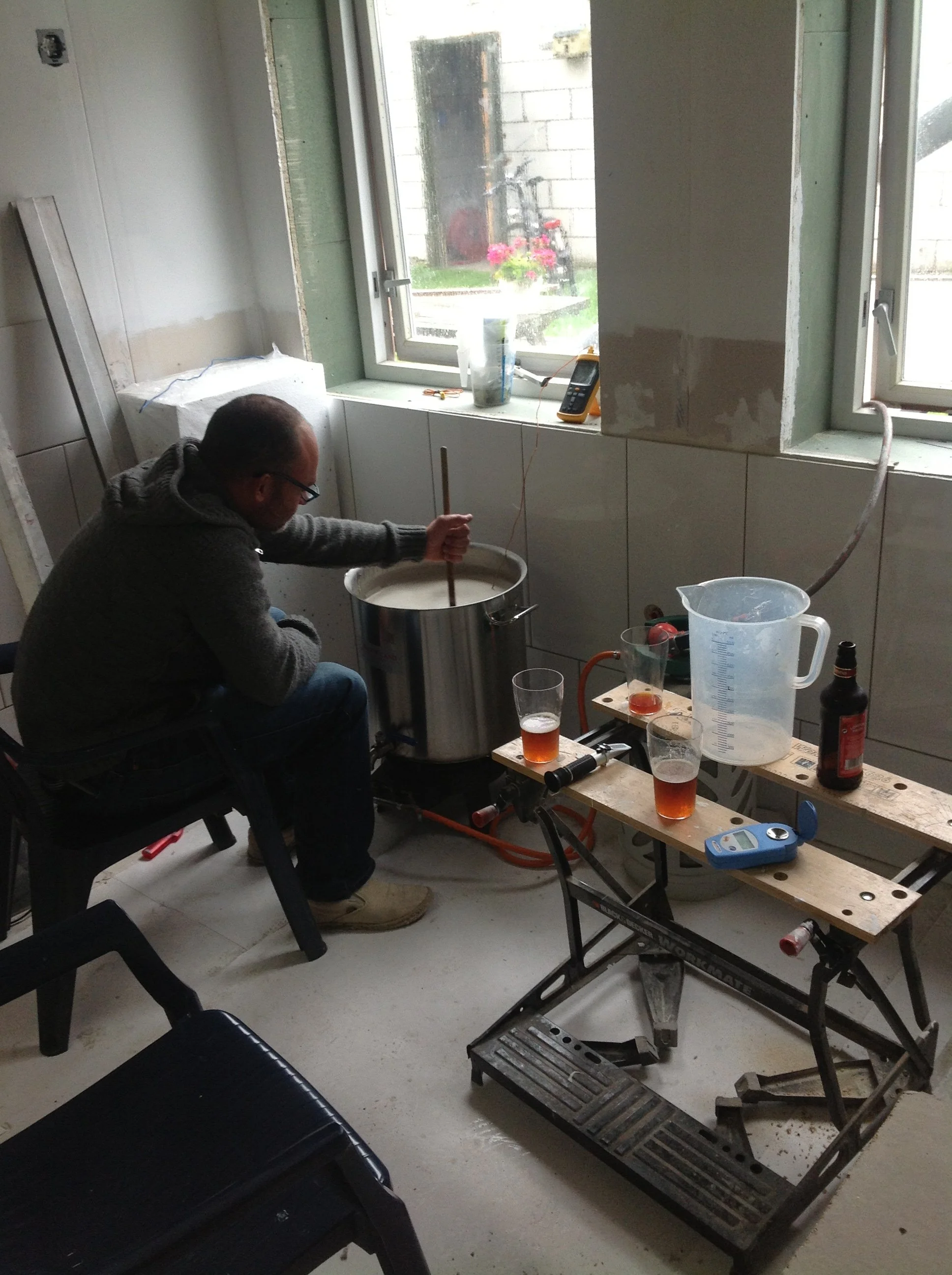 Een man mengt een vloeistof in een grote metalen ketel in een kamer die wordt gerenoveerd, met een raam dat uitzicht biedt op een tuin met bloemen en een fiets.