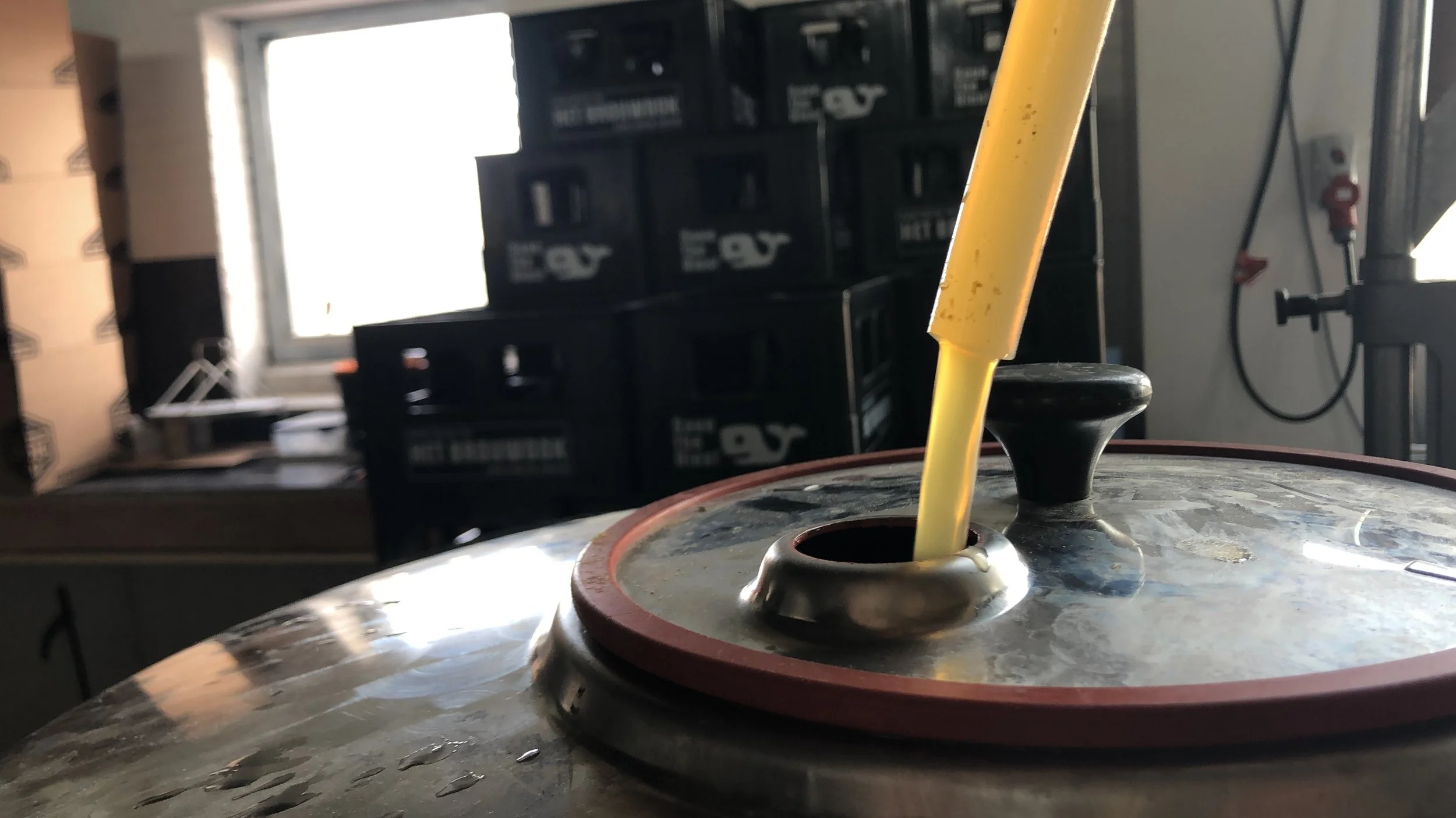 Close-up van een industrieel vat met gele pijp die uit deksel steekt, op de achtergrond zwarte bakken met gereedschap, in een werkplaats of industrieel omgeving.