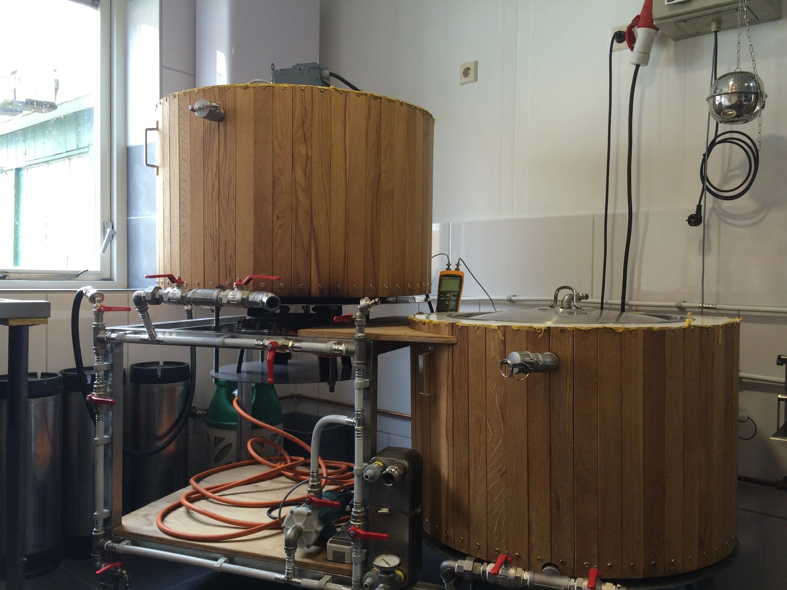 Binnenzicht van een laboratoriumopstelling met een houten vat, buizen, pompen, een multimeter, en elektrische apparatuur, naast een raam.