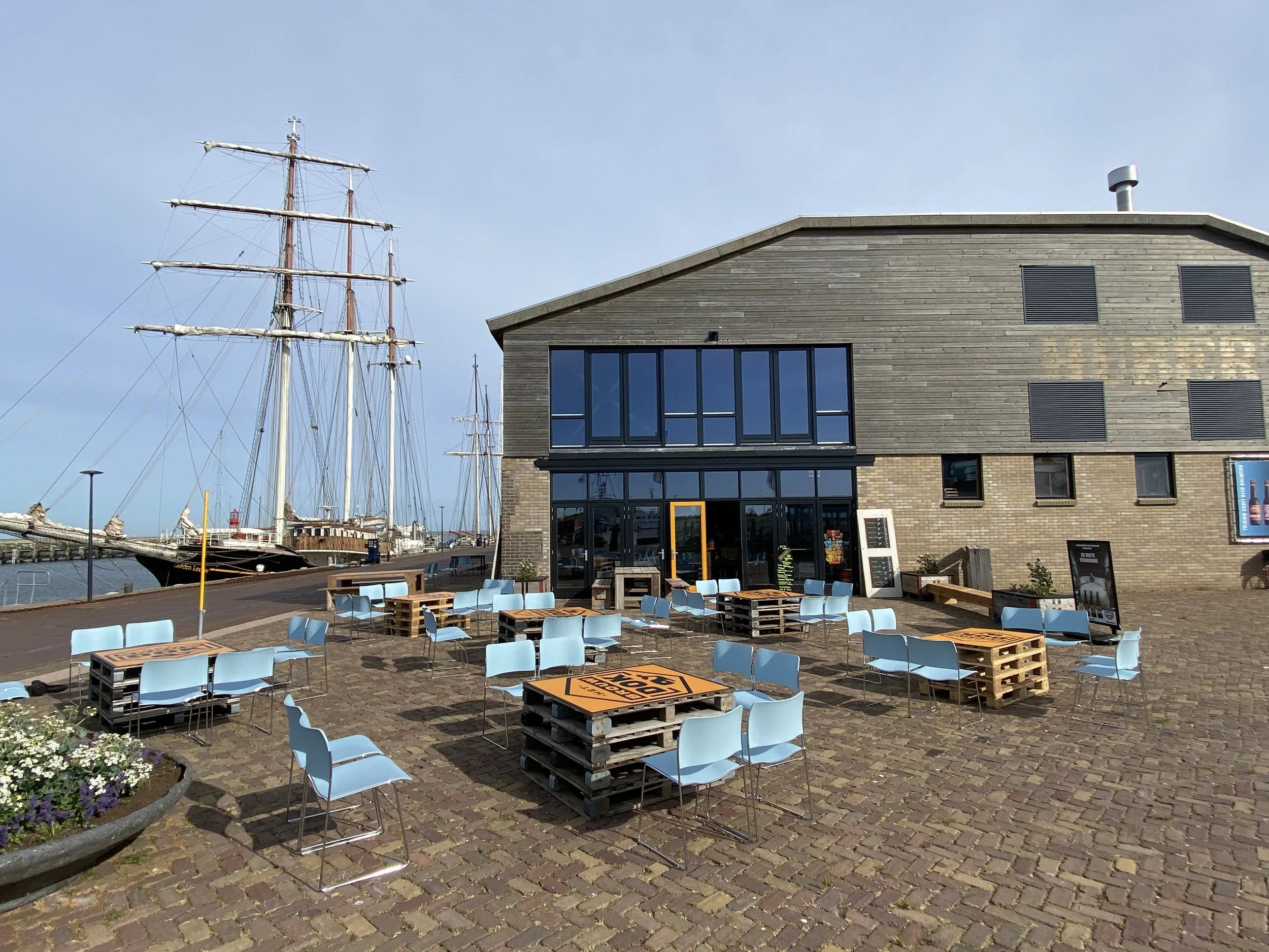 Buiten terras met stoelen en tafels, nabij een haven met schepen, en een modern houten gebouw met grote ramen.