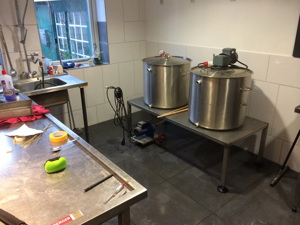  Keuken met twee grote metalen ketels op een tafel, er zijn verschillende gereedschappen en schoonmaakmiddelen op het aanrecht en tafel.