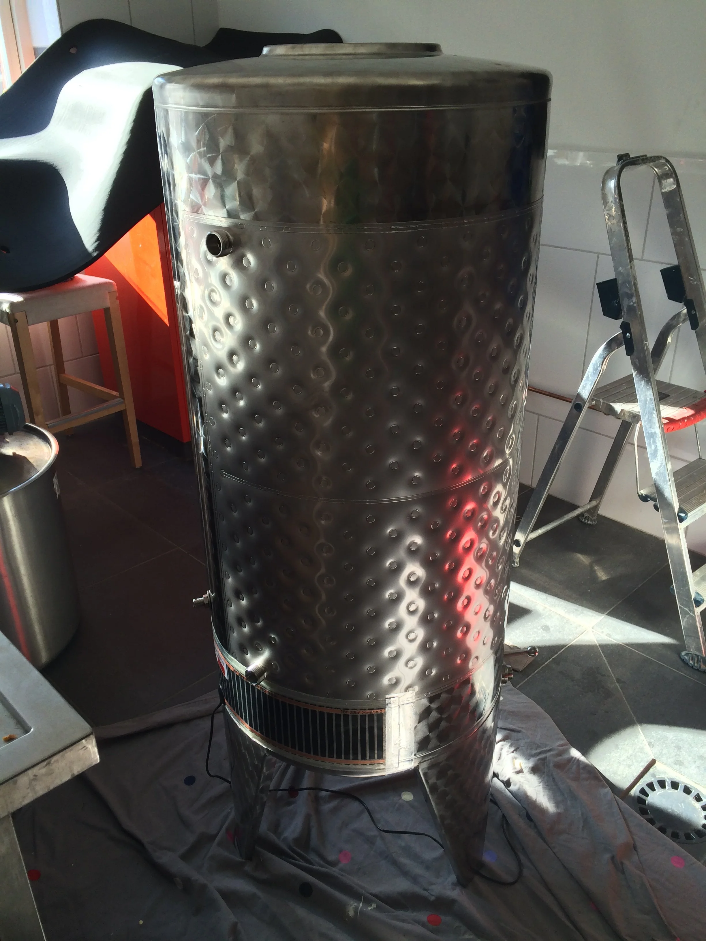 Een rvs-tonnen waterverwarming boiler in een keuken of fabriek, met een ladder ernaast en een zwarte doek onder de boiler.