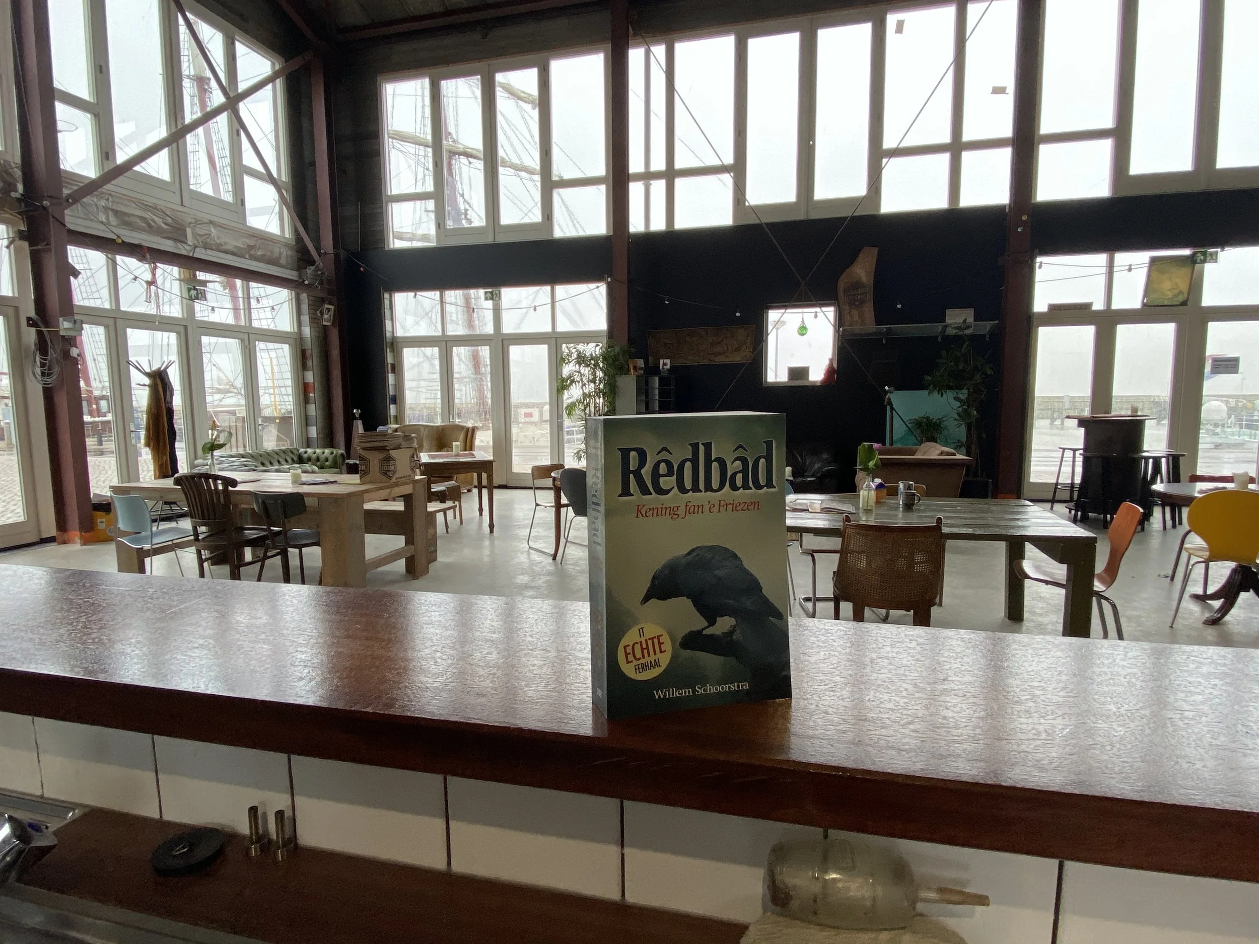 Interieur van een café of restaurant met grote ramen, diverse stoelen en tafels, en een promotiebord voor een boek op de toonbank.