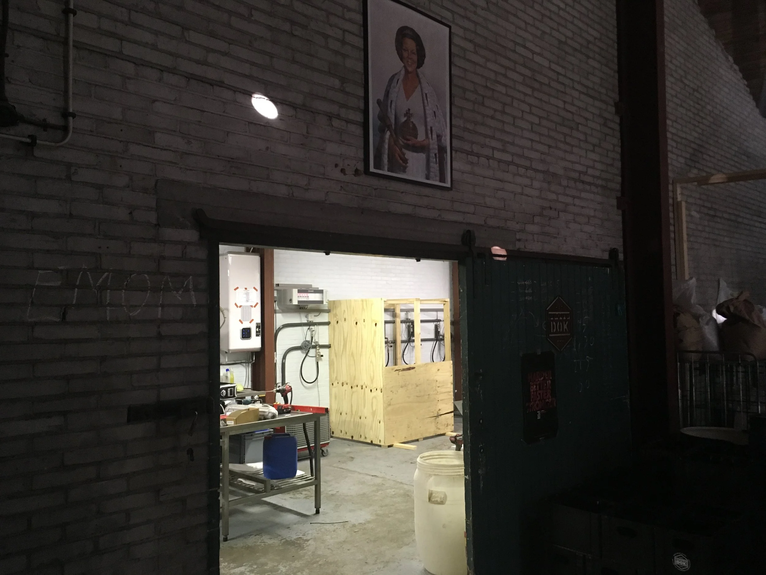 Binnenzicht van een werkplaats of garage met een betonnen vloer en bakstenen muur. In het midden is een houten structuur of bar nog in opbouw. Er hangen wat gereedschappen en keukenspullen aan de muur, en er zijn diverse gereedschappen en dozen op ee