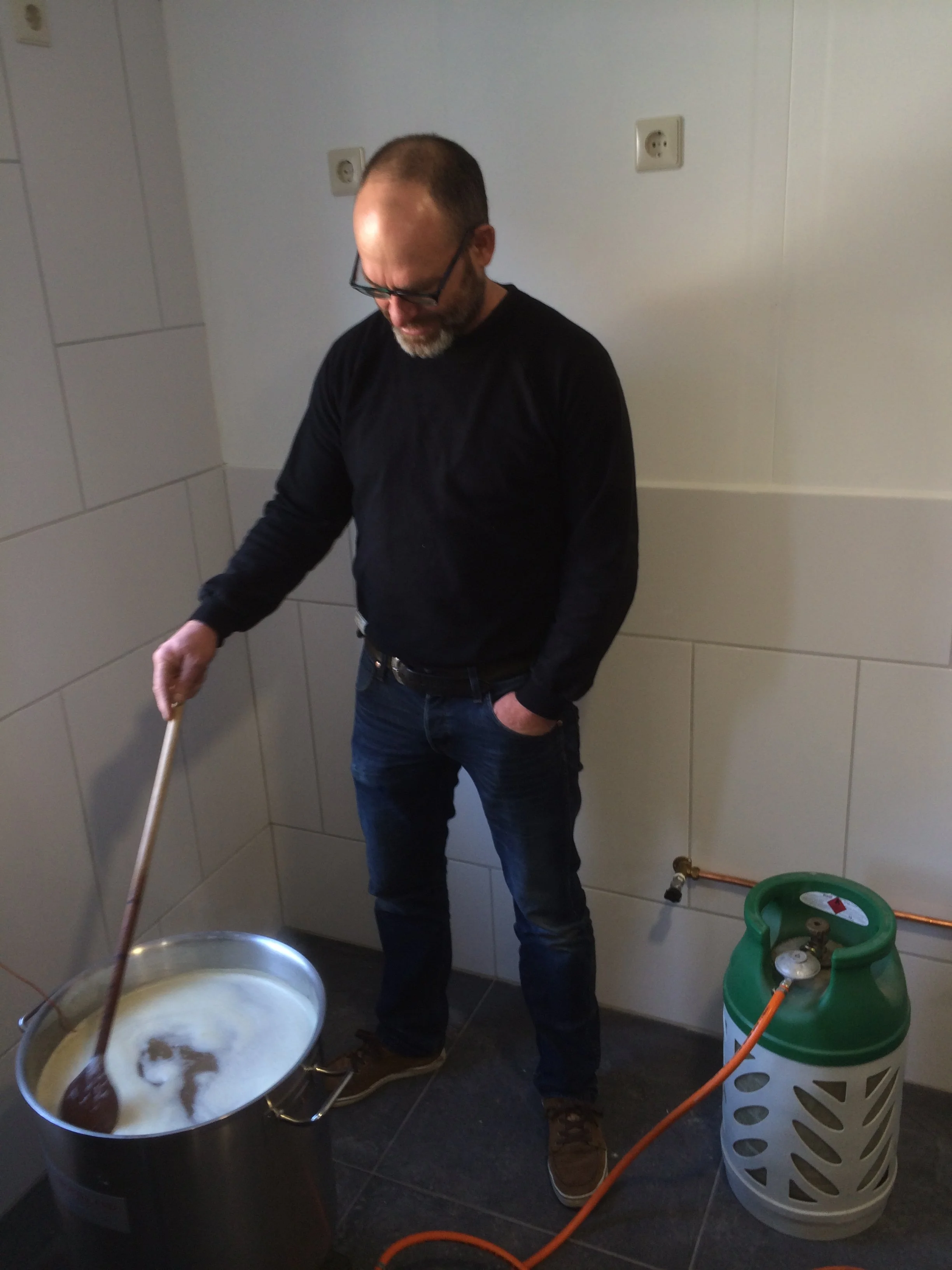 Man in een zwarte trui en blauwe jeans die roert in een grote steelpan met schuim en vloeistof in een keuken, met gasfles en slang ernaast.