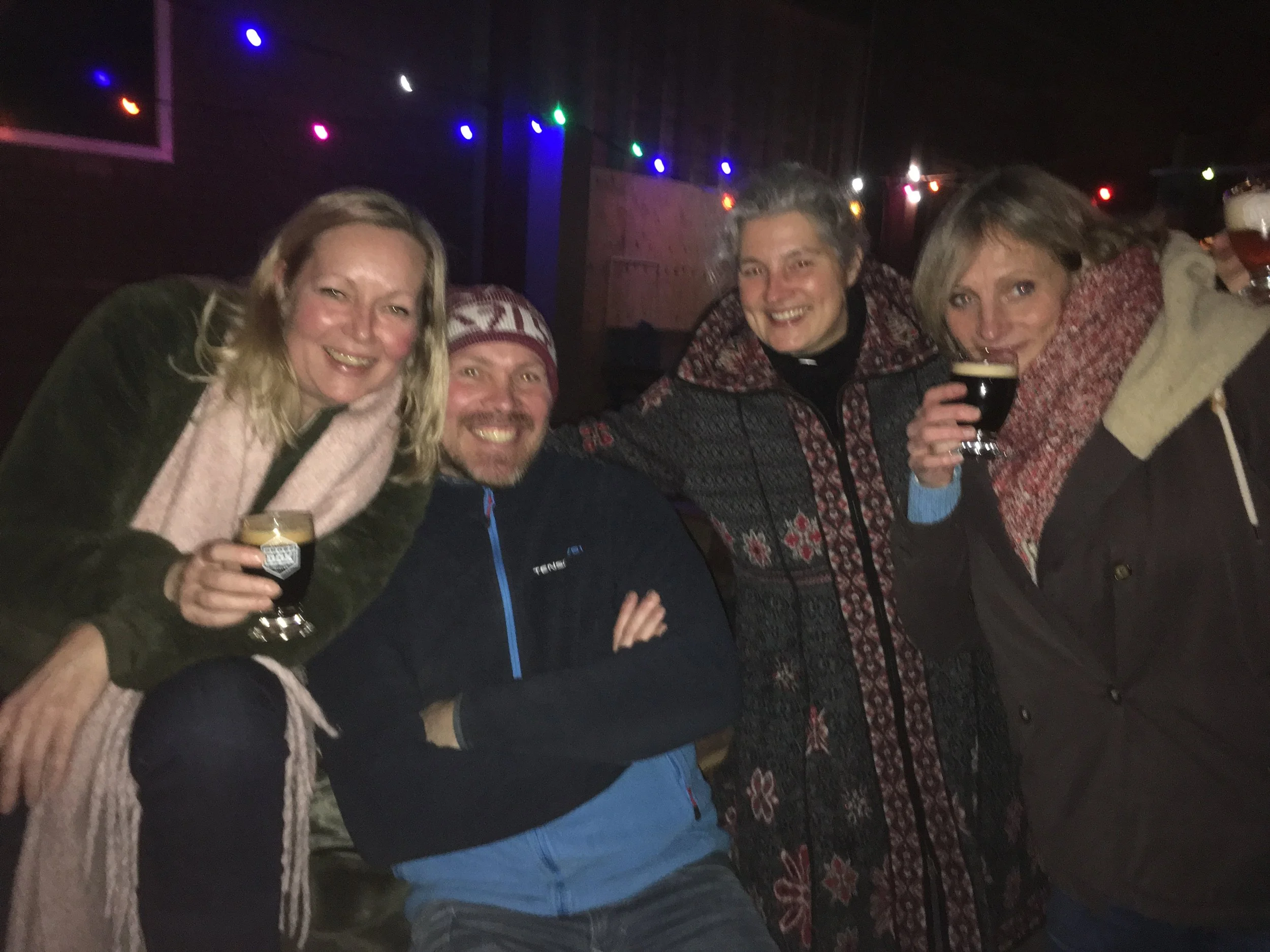 Vijf mensen die plezier hebben tijdens een feestje, met kleurrijke kerstverlichting op de achtergrond, ze drinken samen en lachen.
