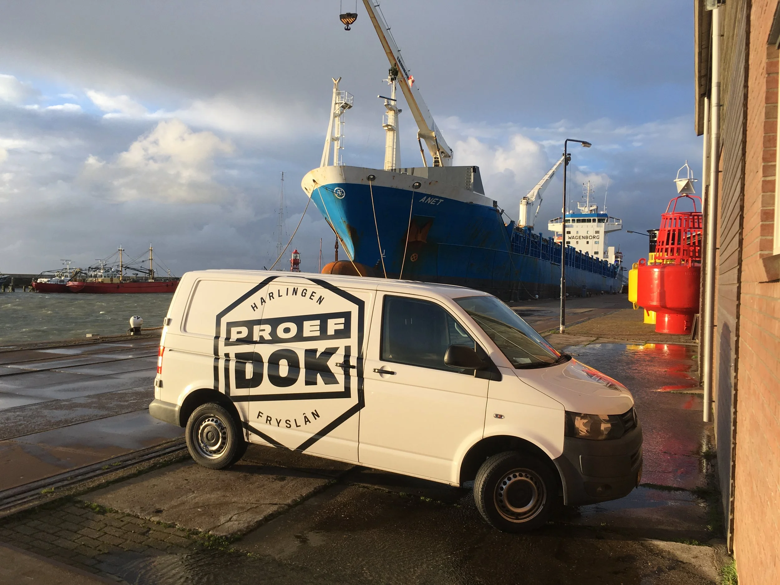 Een grote blauwe vrachtboot ligt in de haven, met kranen erboven. Op de voorgrond staat een witte bestelbus met het logo 'PROEFFRYSLAN'.