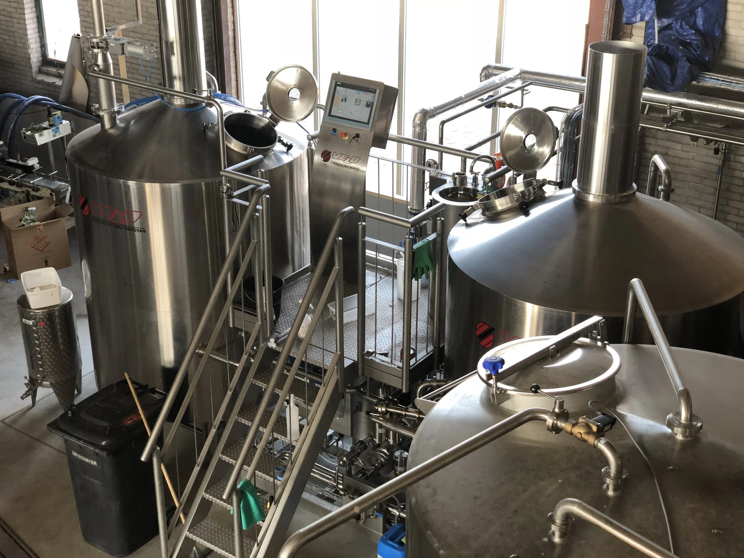 Stainless steel fermenters en apparatuur in een brouwerij, met een bedieningspaneel en metalen piping.