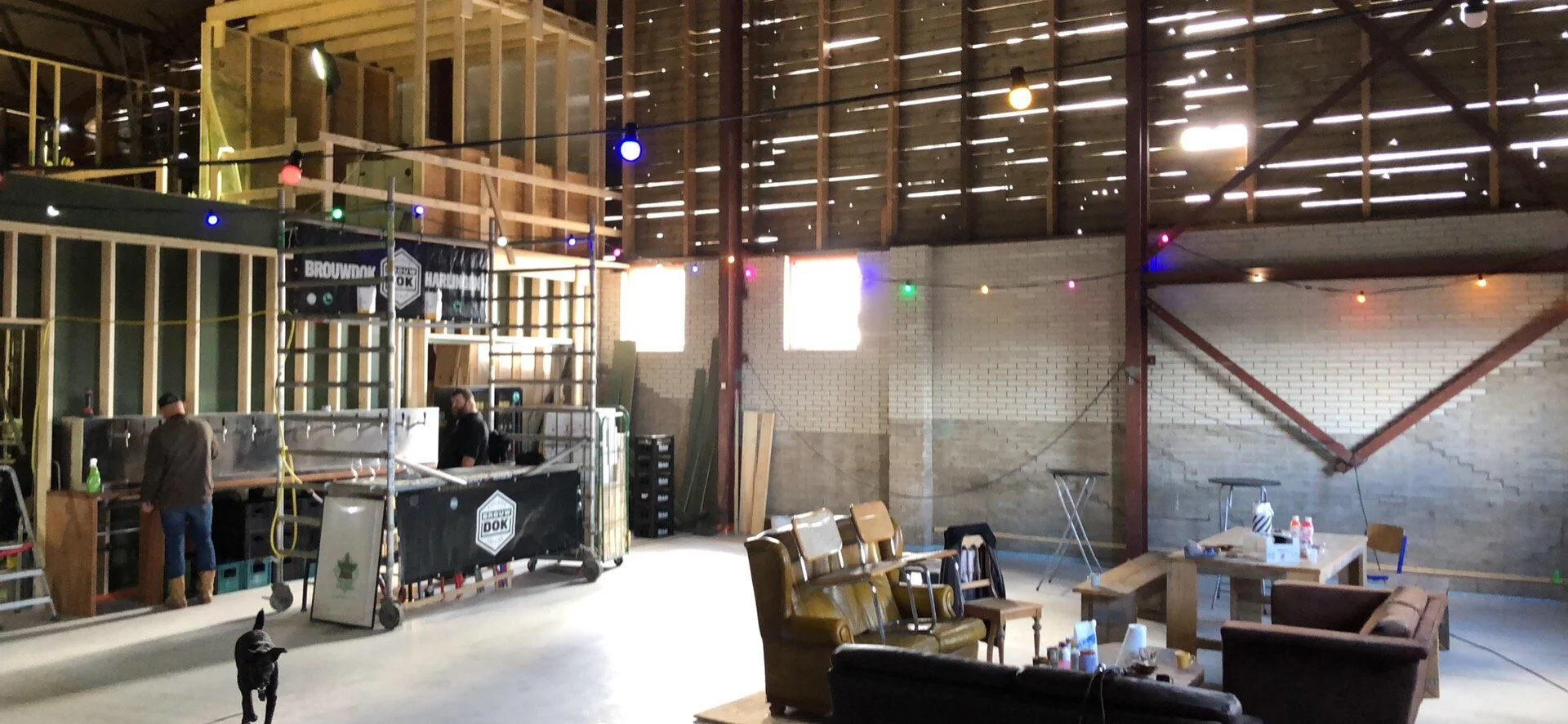 Binnenkant van een een grote, ongelakte houten bouwschuur met tijdelijke verlichting en meubels, inclusief banken en stoelen, en een paar mensen die werken aan de bouw of het opzetten van een evenement.