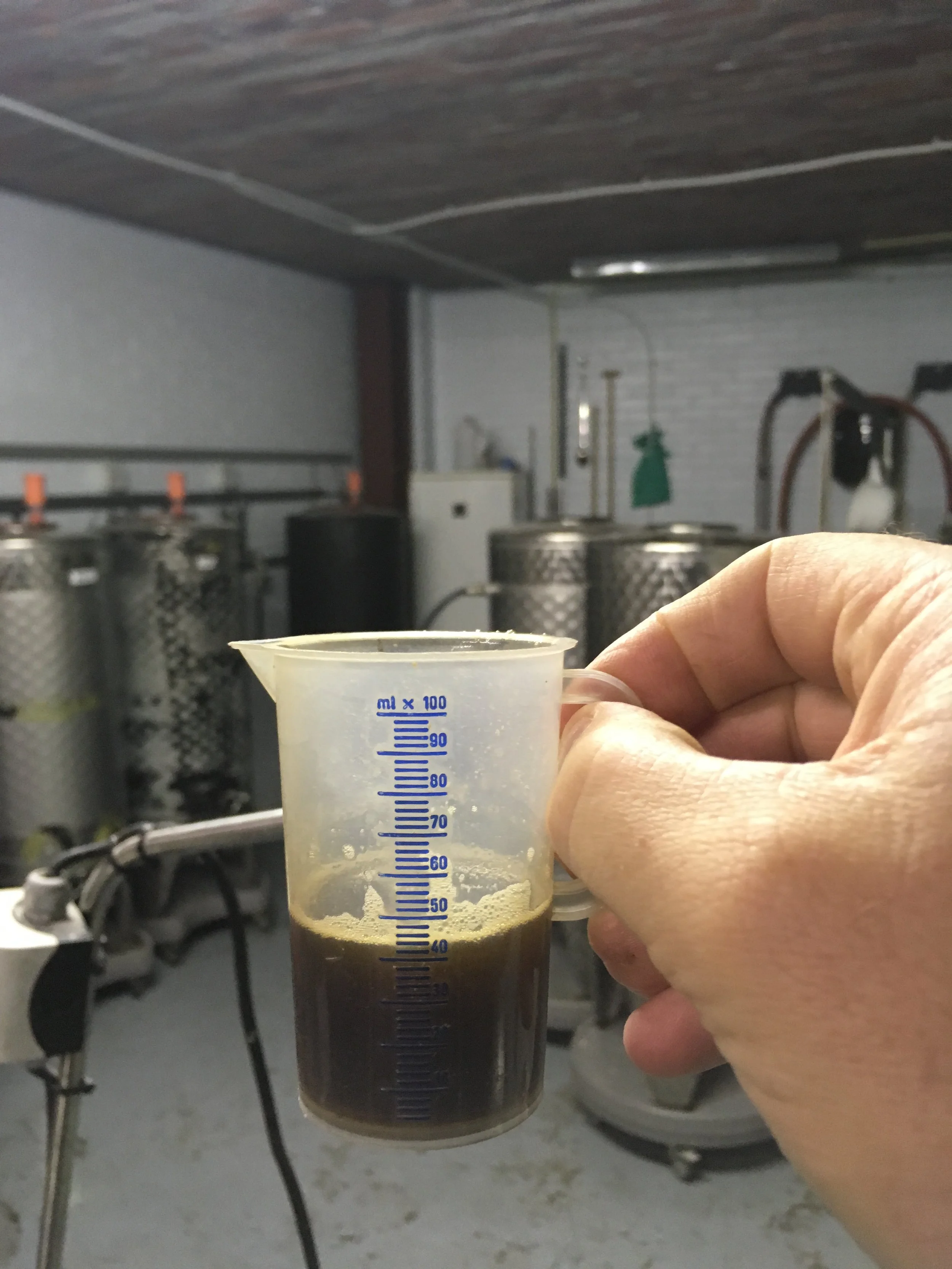 Een hand die een plastic meetbekertje vasthoudt met een donkergekleurde drank erin, mogelijk koffie, espresso of Cold brew, in een professionele keuken of brouwerij, met metalen tanks en apparatuur op de achtergrond.