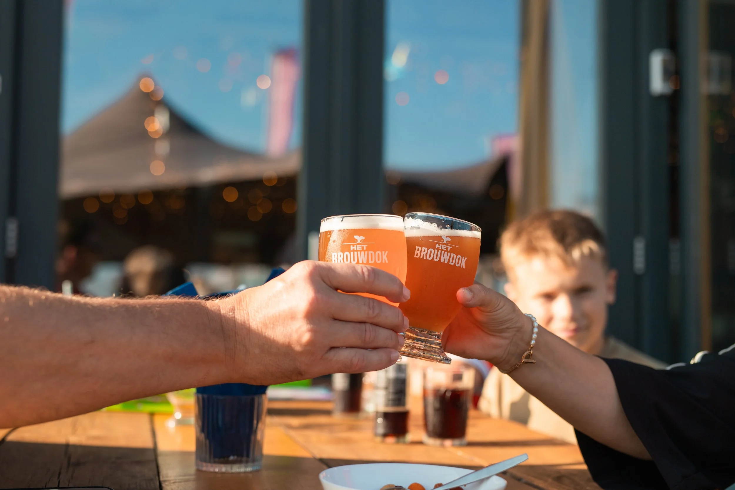 Twee mensen heffen glazen met een lichte amberkleurige drank in een cirkel in een feestelijke toast, op een houten tafel met drinken en een wit bord, buitenshuis bij daglicht.