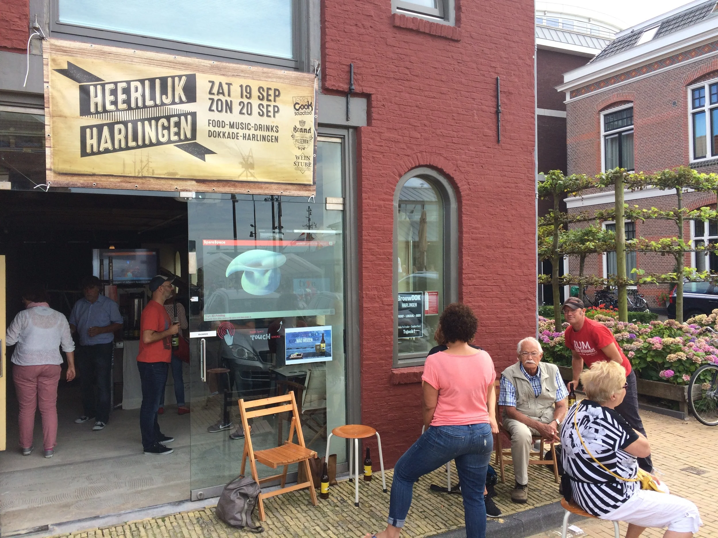 Mensen verzamelen zich buiten een restaurant of café met rode bakstenen gevel en grote ramen, waar een grote gele banner hangt met de tekst 'Heerlijk Harlingen' en data van een evenement. Er zijn tafels, stoelen en bloemen in de omgeving, en bezoeker