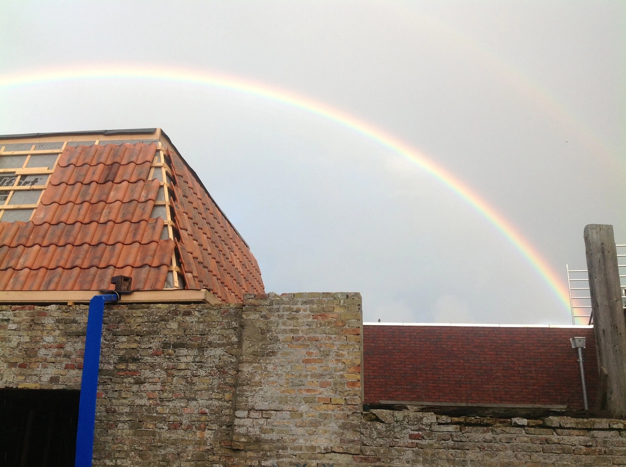 Een regenboog boven een dak met rode dakpannen en een stenen muur.