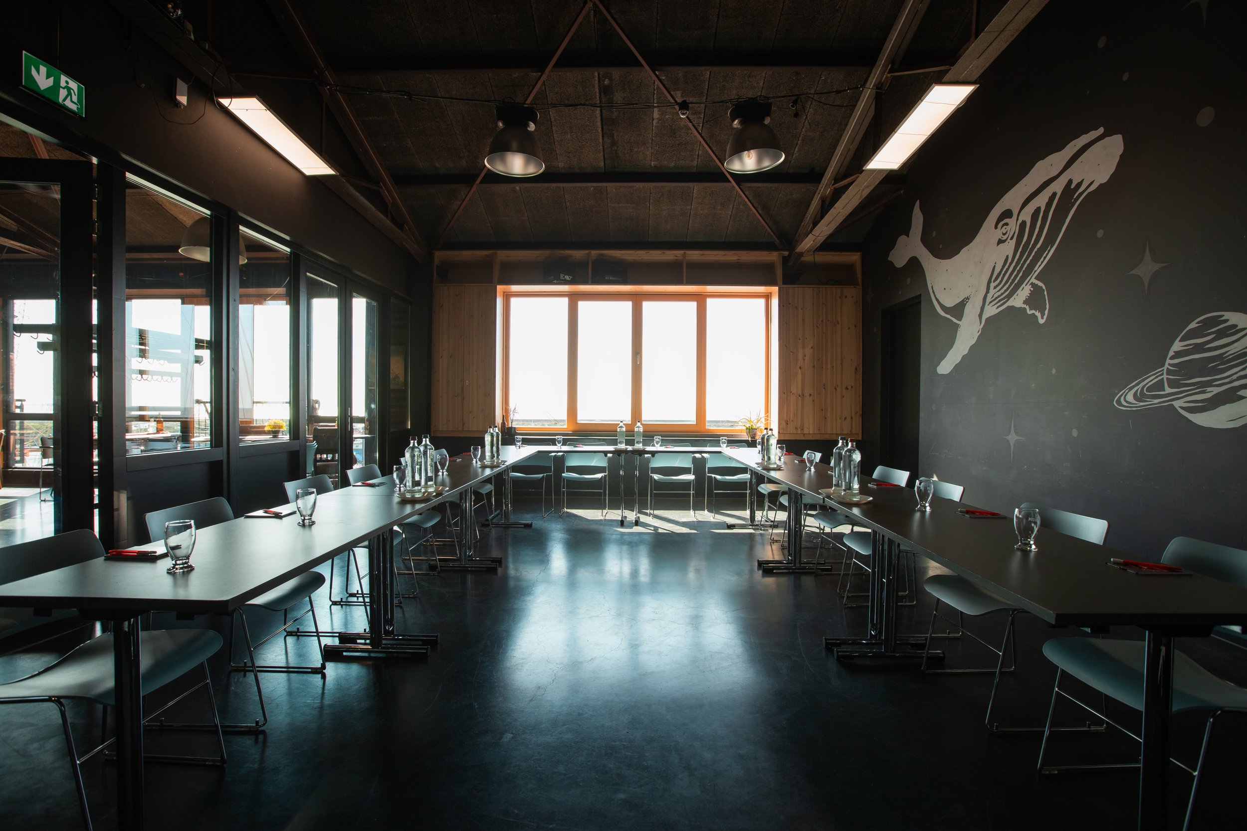 Een vergaderruimte met lange tafels en stoelen, grote ramen die licht binnenlaten, een muur met een ruimte-achtige muurschildering, en een plafond met industriële verlichting.