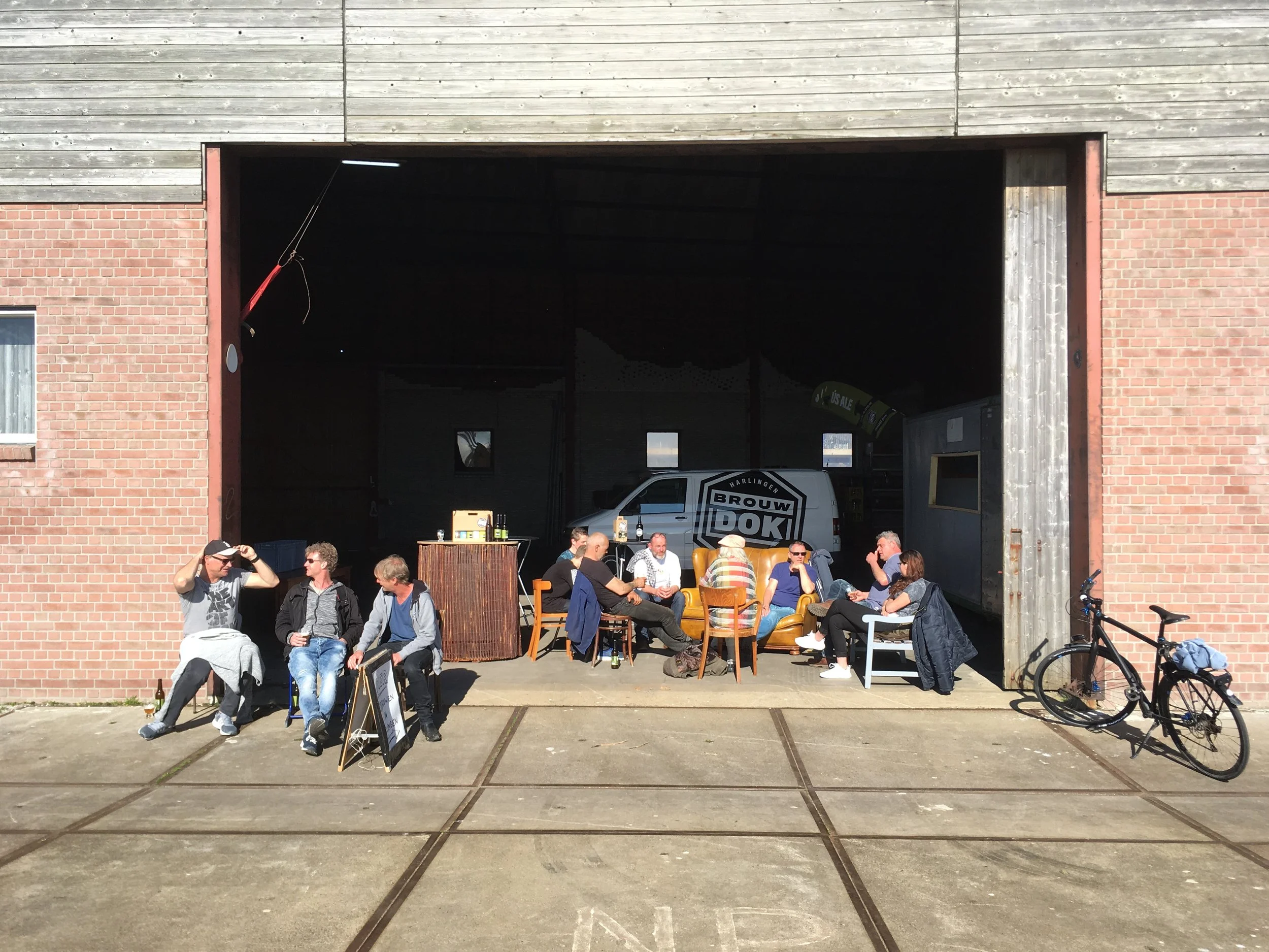 Buitenevenement met mensen die op straat genieten, met tafels, stoelen en een brew van auto's in de voorkant, onder een grote houten en bakstenen gevel.