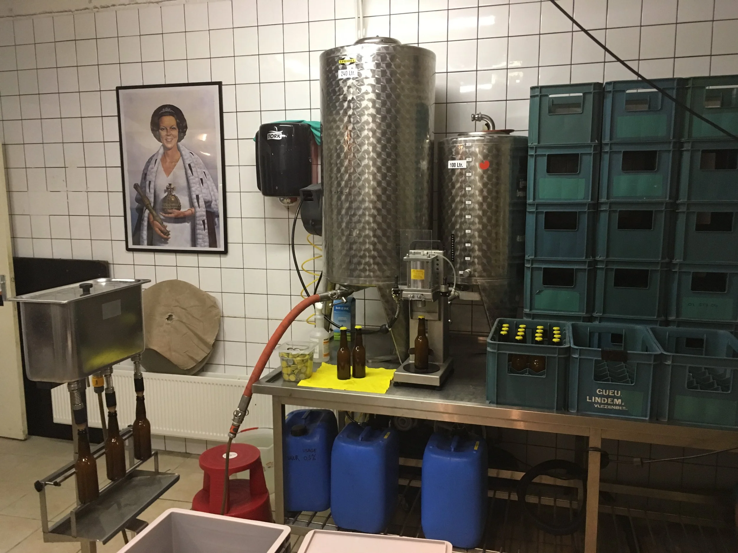 Diverse drankflesjes en containers, een poster van een vrouw met een kroon, en stapelbare groene bakken in een ruimte met witte betegelde muur.