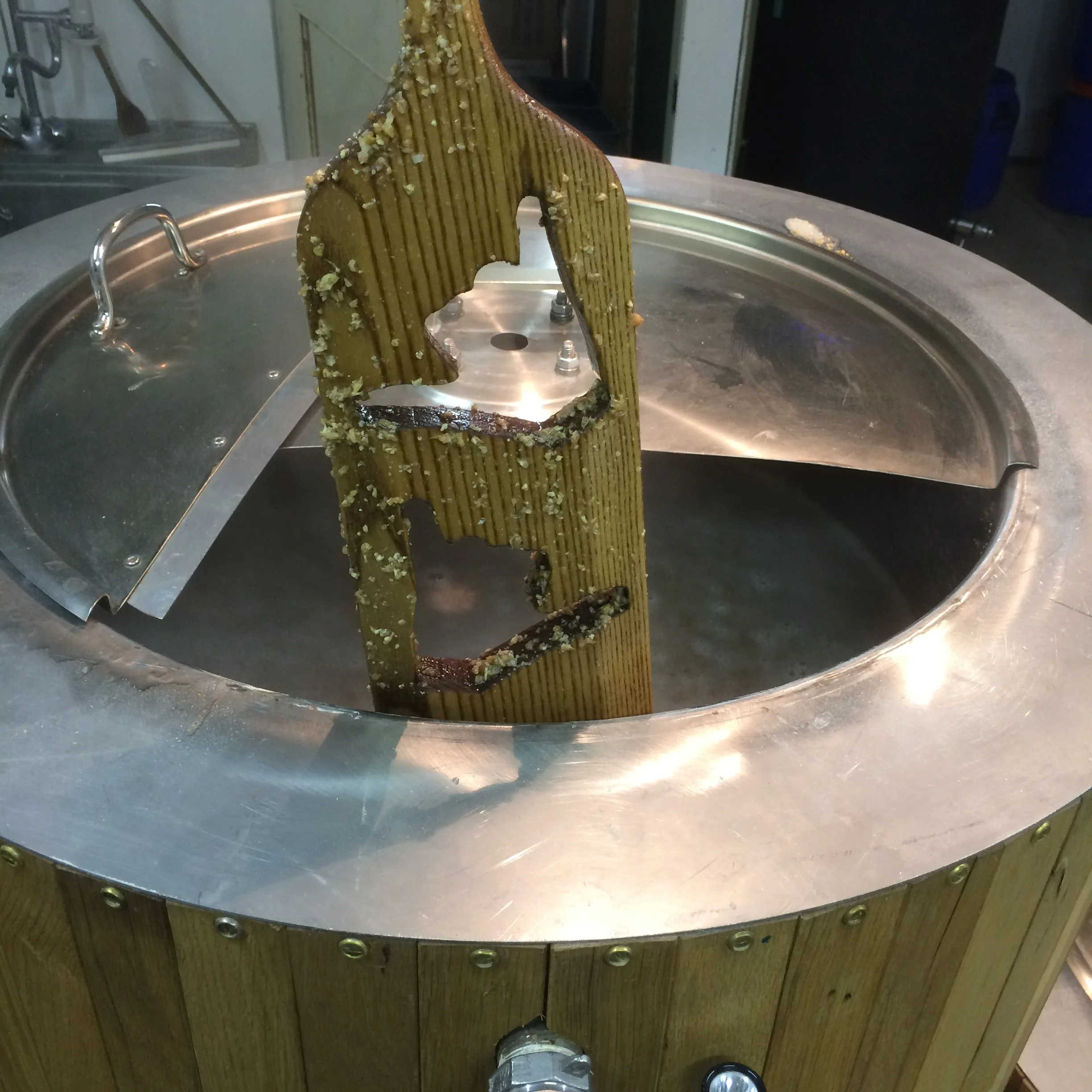 Een grote houten structuur met een halve cirkelvorm en verticale groeven, voorzien van kleine stukjes knetterschaam en een uitgesneden vorm, bevestigd op een ronde metalen plaat. Op de achtergrond is een keuken te zien.