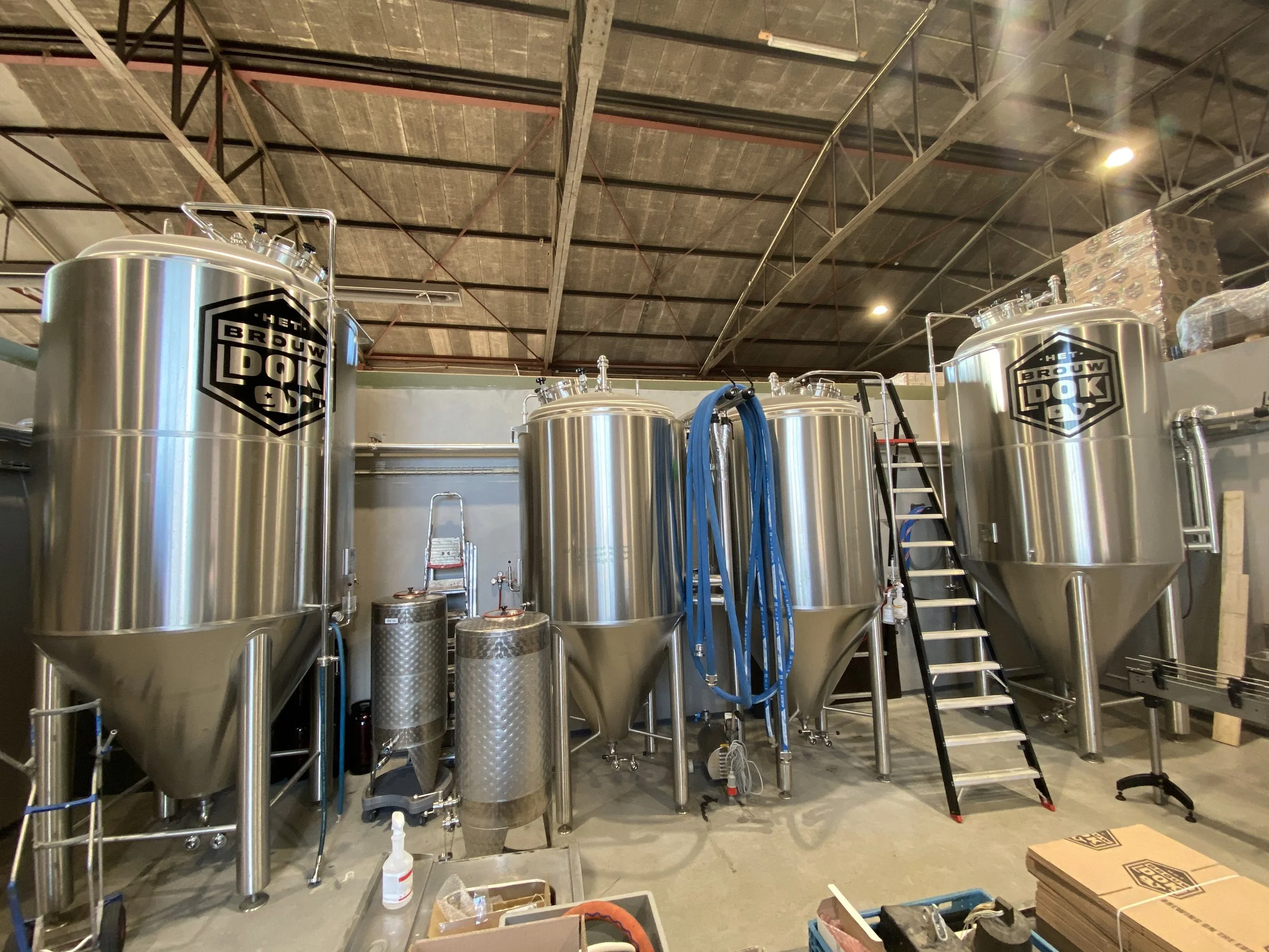 Rvs brewing tanks met het logo 'Het Brouwdok' binnen in een fabriek of brouwerij, met allerlei apparatuur, ladders, en verpakkingsmaterialen zichtbaar.