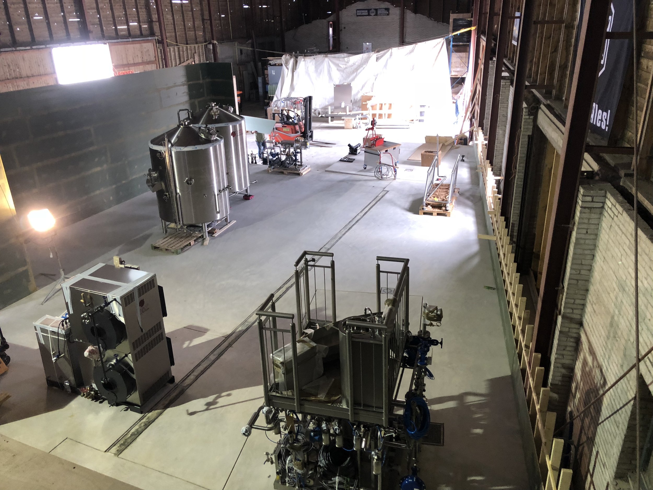 Interne foto van een grote bedrijfsloods of brouwzaal met apparatuur, werkplatforms, en materialen voor bierproductie.