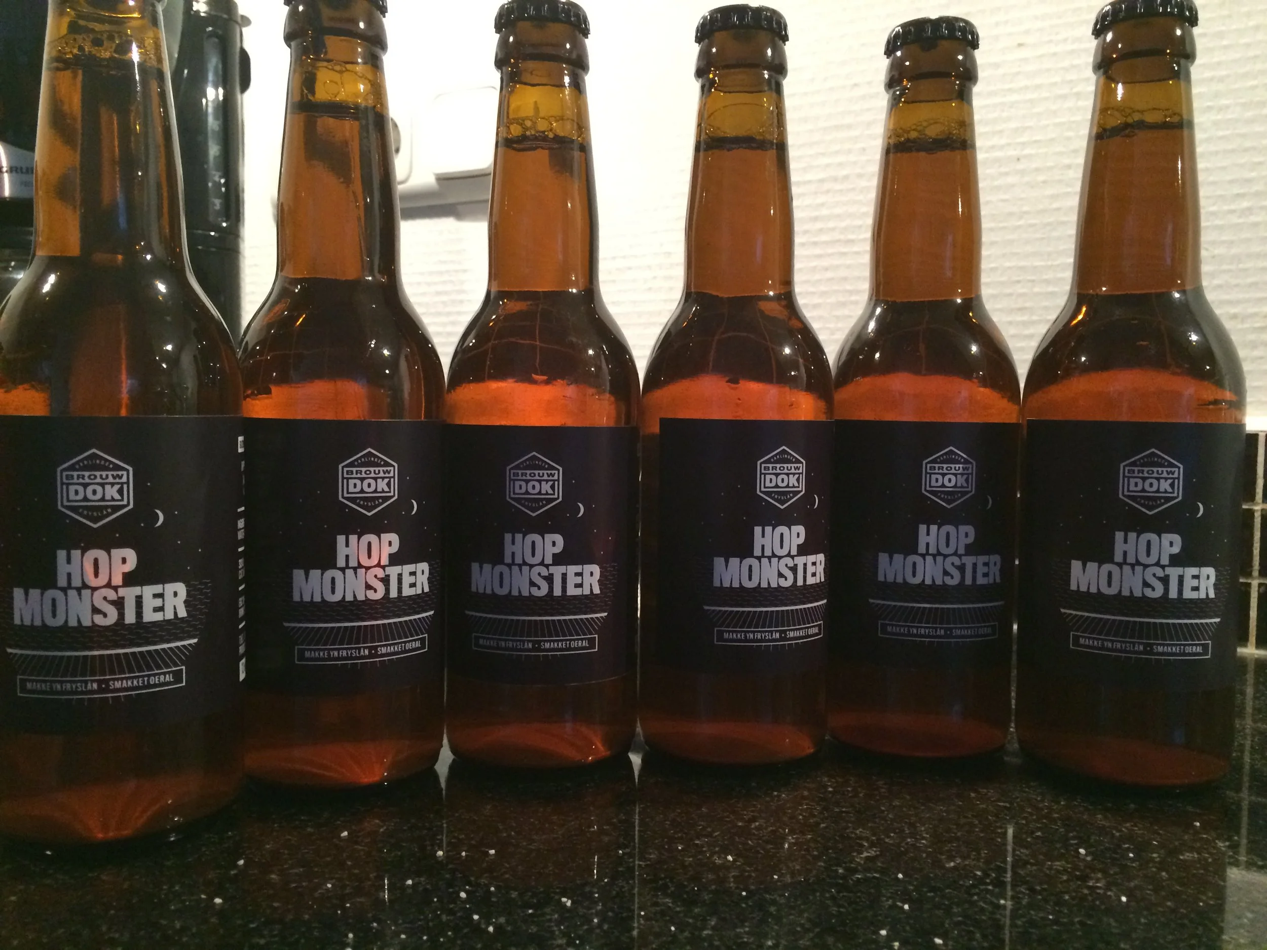 Zes bierflesjes met zwarte labels en de tekst 'Hop Monster', geplaatst op een zwarte tafel. Op de achtergrond zijn witgekleurde muur en elektrische apparaten zichtbaar.