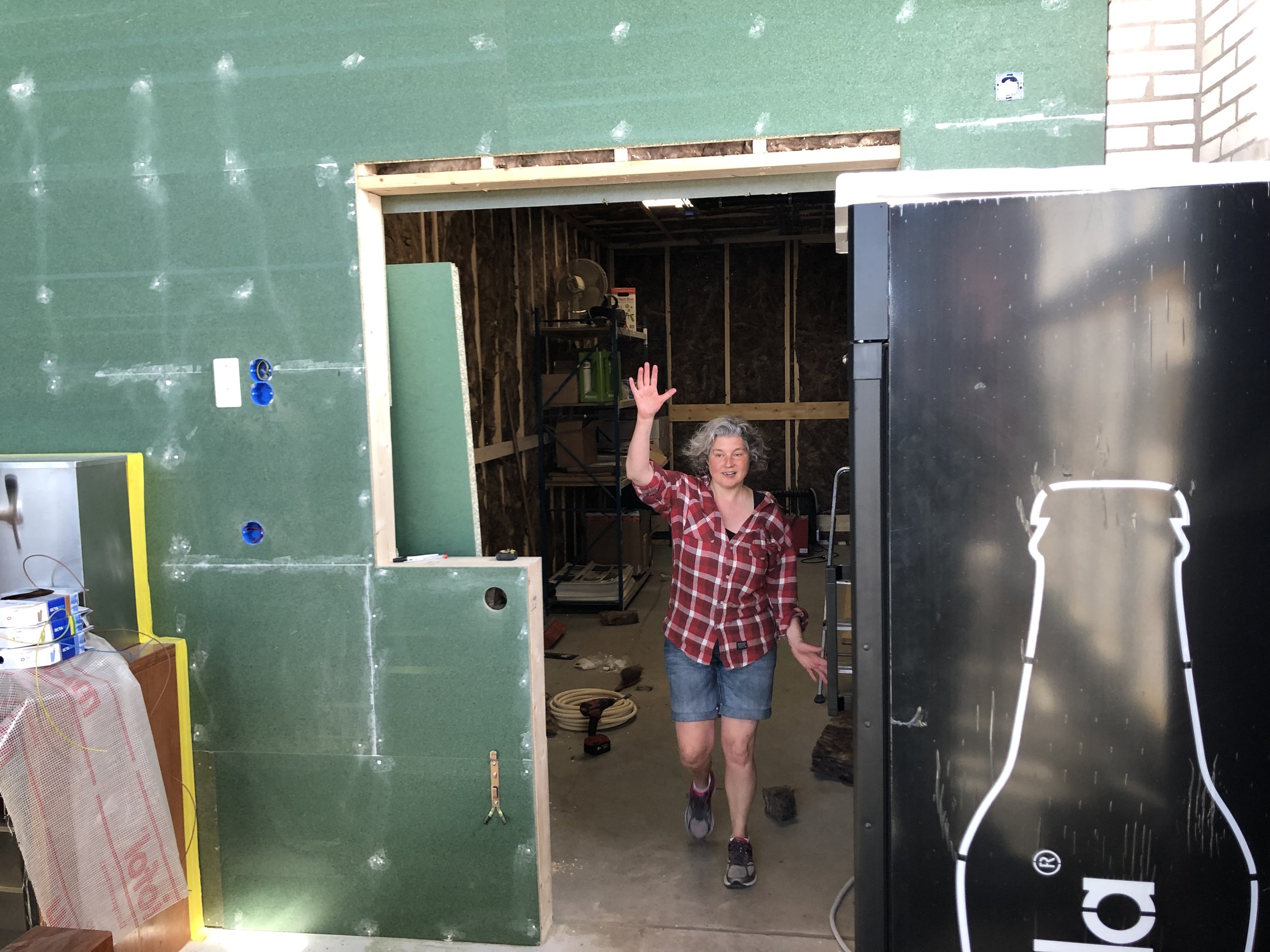 Vrouw in casual kleding en schoenen staat in een bouwplaats, zwaait en glimlacht naar de camera. De achtergrond toont een kamer in aanbouw met houten frames en gereedschap.