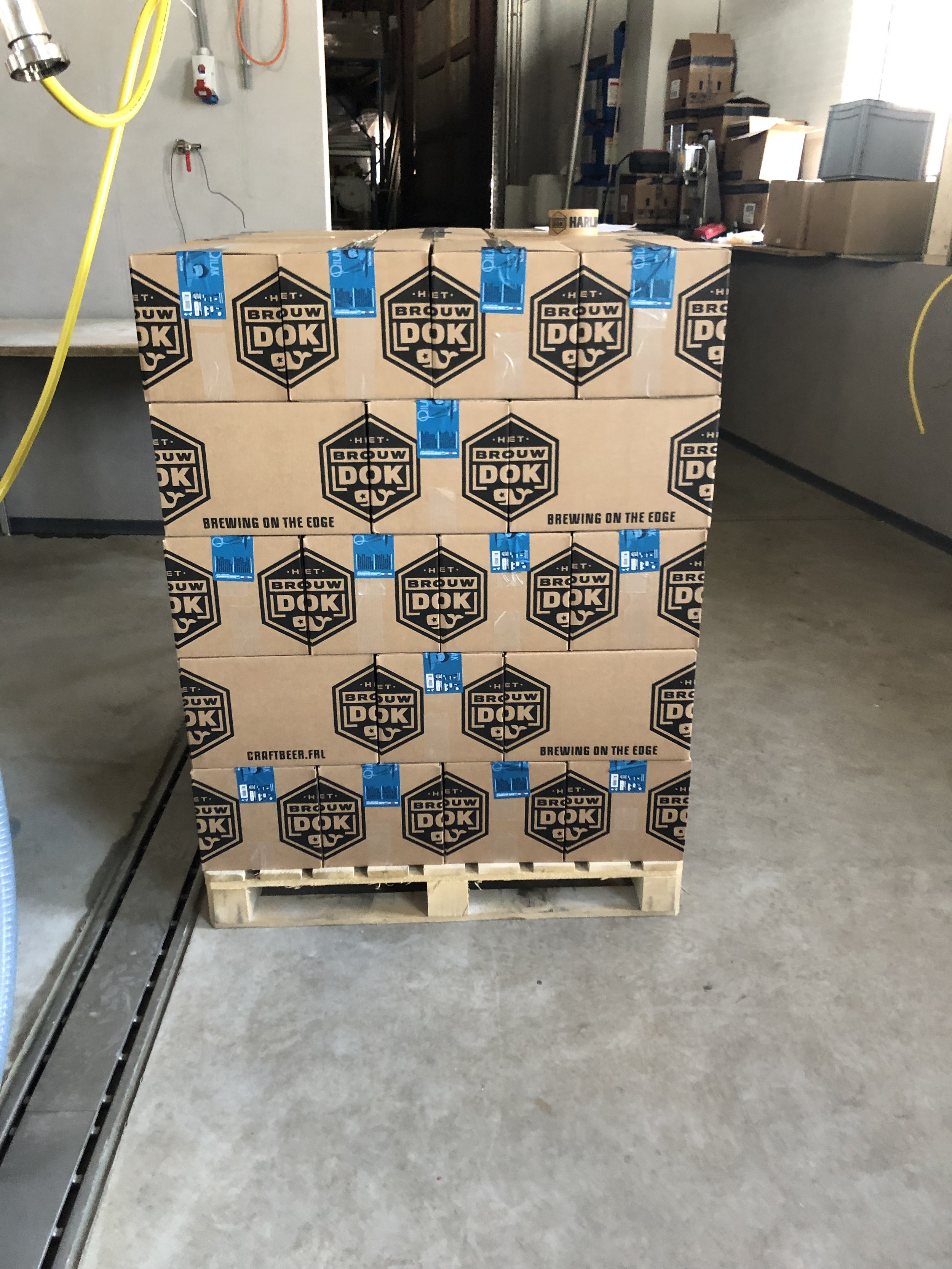 Stapel kartonnen dozen met het logo 'HET BROW DOK' en de slogan 'Brewing on the Edge', geplaatst op een pallet in een magazijn of opslagruimte.