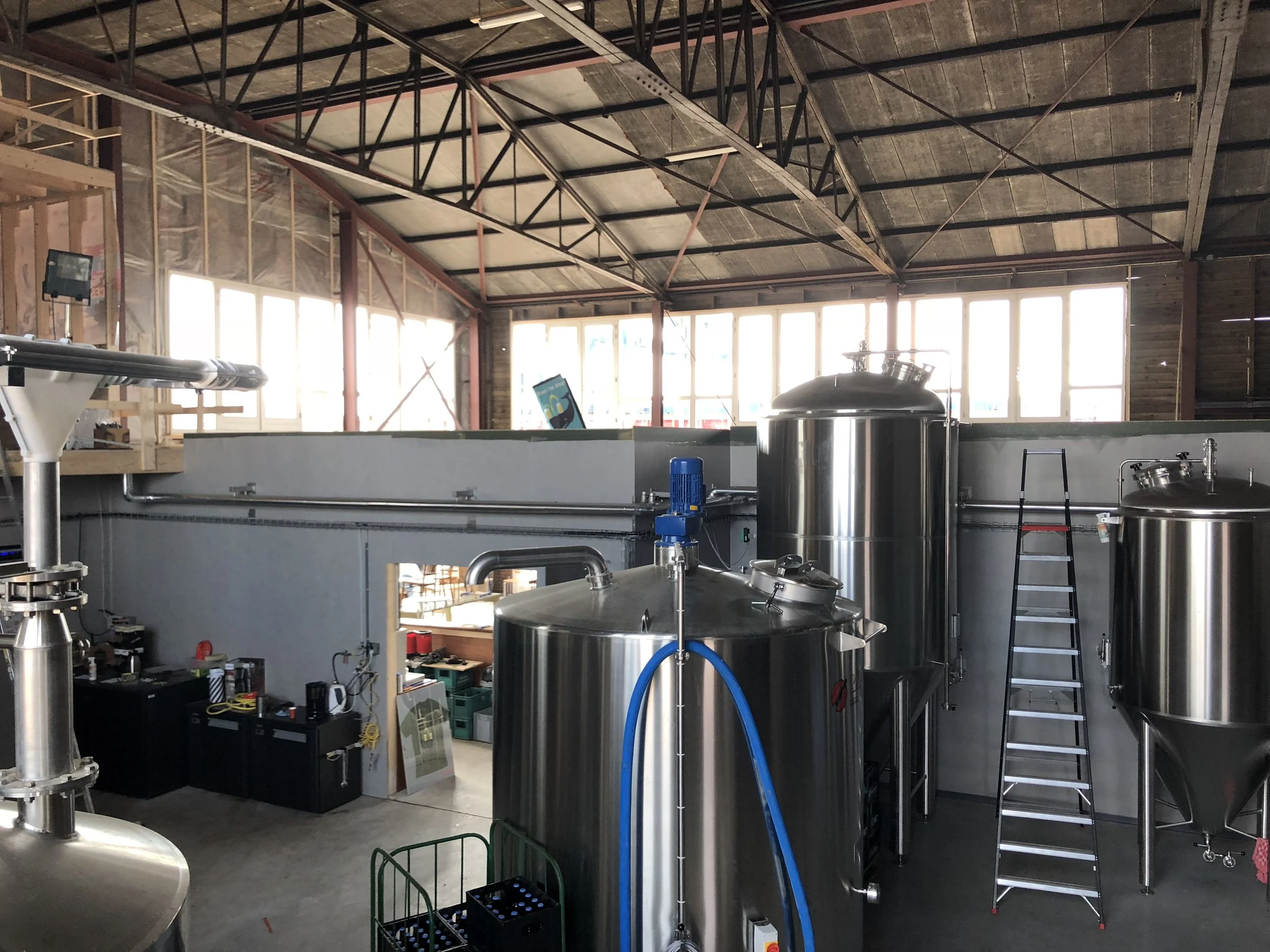 Industriële ruimte met grote roestvrijstalen tanks, een ladder, en machines, mogelijk voor brouwerij of productie, met grote ramen aan de achterzijde.