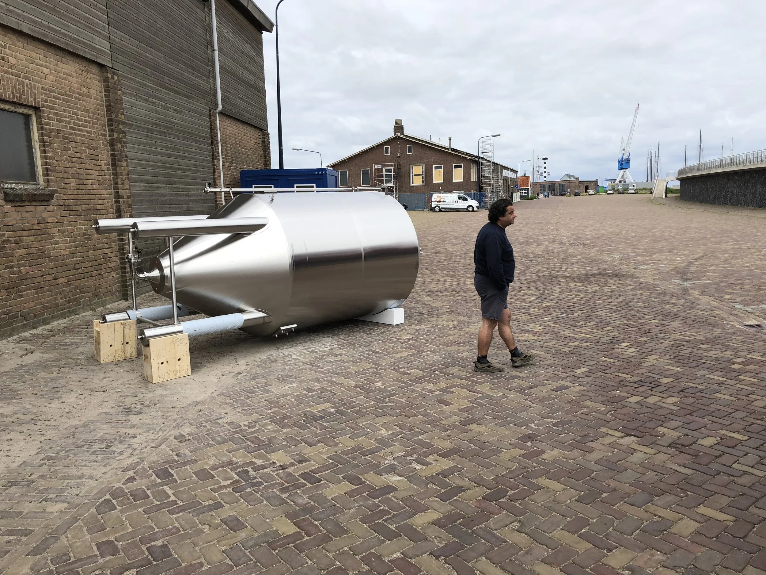 Een man wandelt op een stenen promenade bij een haven met schepen en een grote blauwe kraan op de achtergrond.