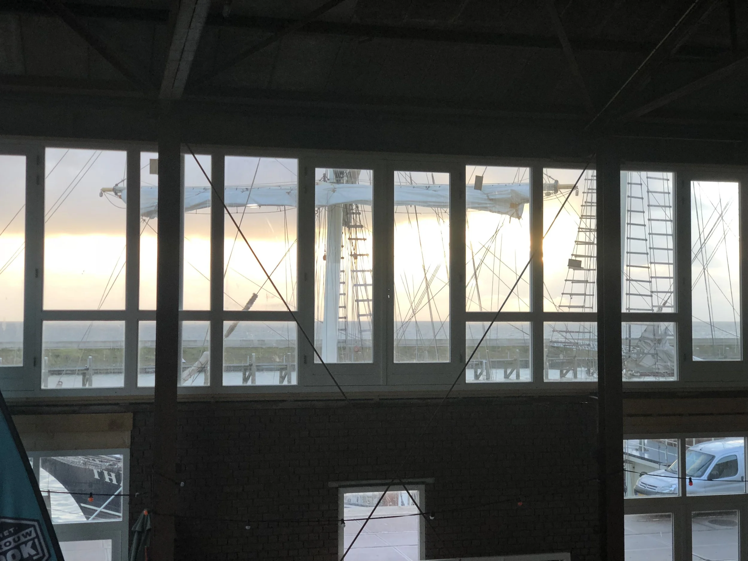 Een foto genomen door een raam van een interieur, met uitzicht op een zeilboot met een kapotte zeil buiten, bij zonsondergang of zonsopgang.