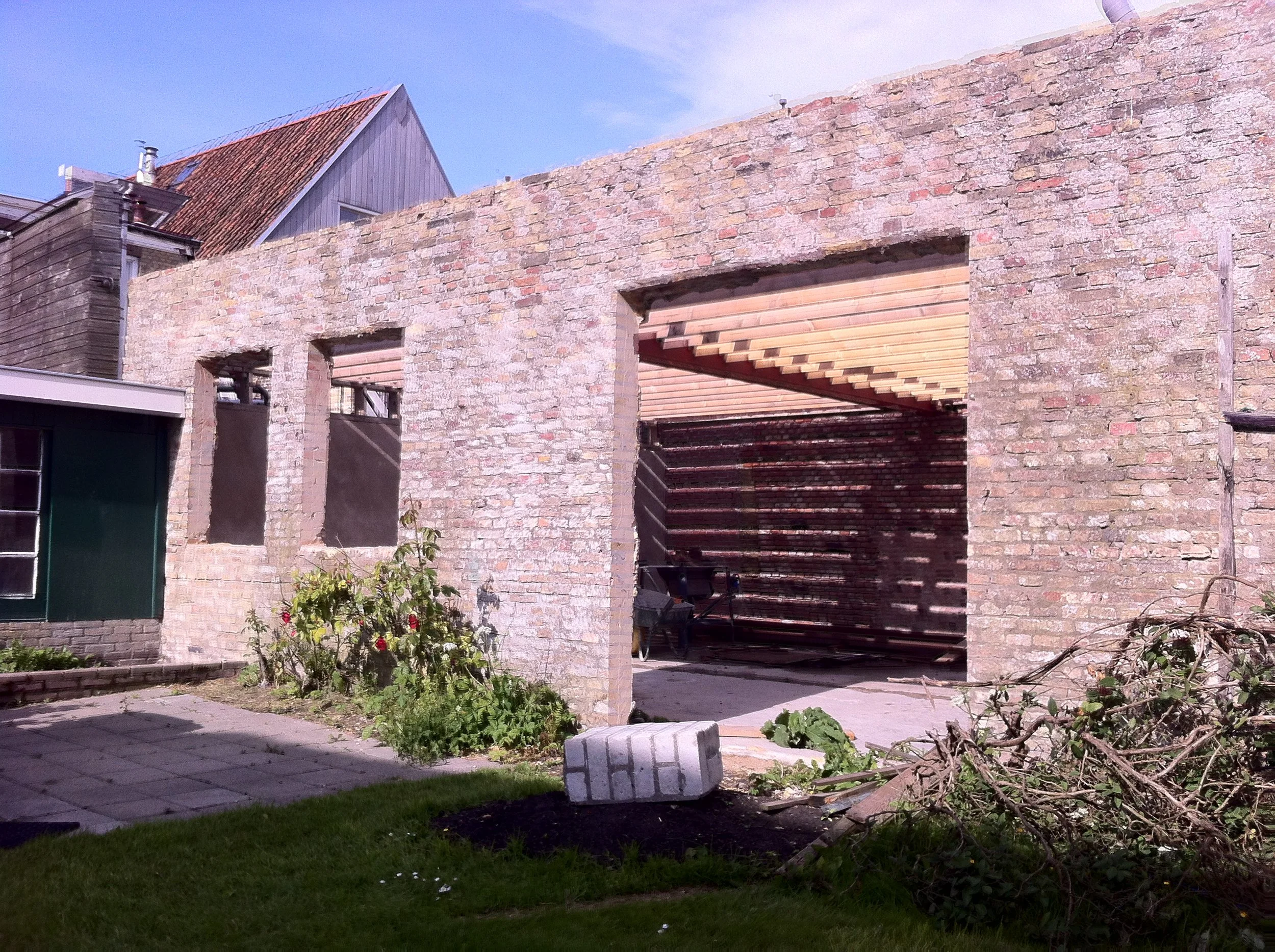 Een bouwplaats met een nog onvoltooide bakstenen muur en een houten constructie erboven, planten en bomen op de voorgrond, en een huis met een puntdak op de achtergrond.